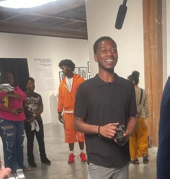 A young man smiling and holding a camera in an art gallery, with other visitors in the background.