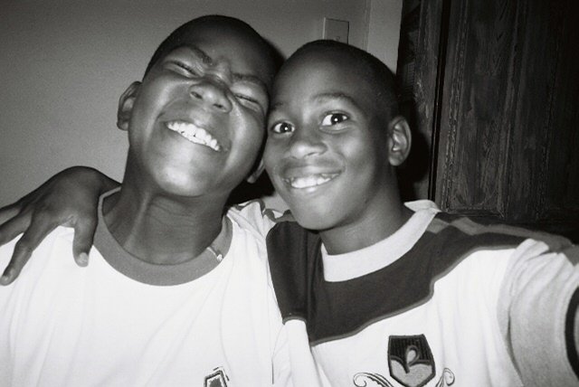 Two young boys smiling and posing for a selfie indoors, with one boy wearing a white shirt and the other in a striped shirt.