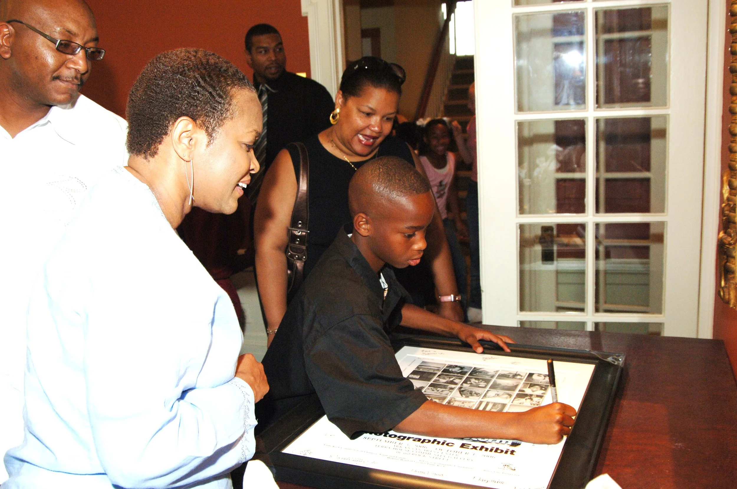 Young boy signing a document or guestbook at a framed display table while women and a man look on, in a room with a glass-paneled door in the background.