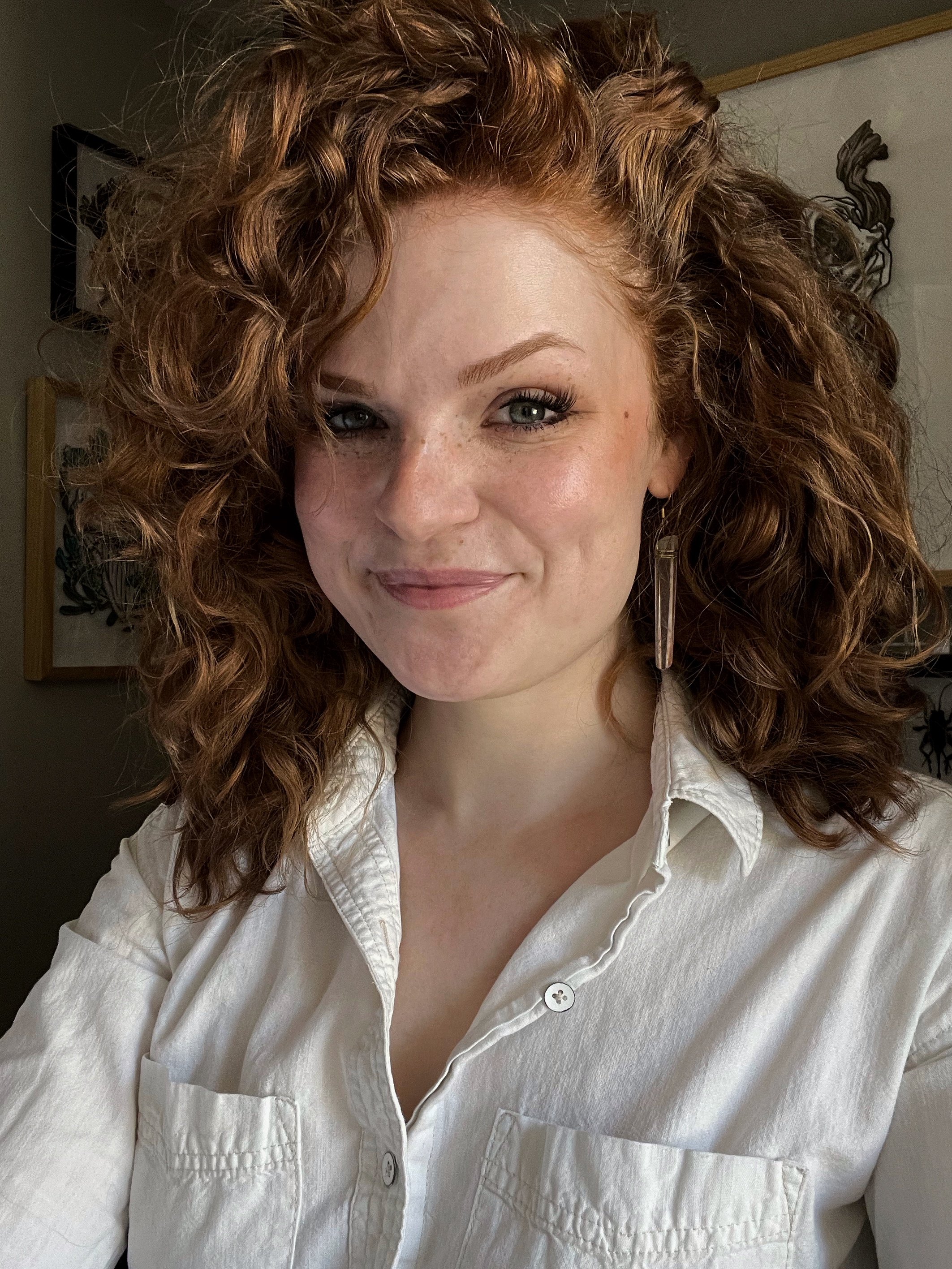 Close-up of a woman with curly red hair, wearing a white button-down shirt and long earring, smiling indoors.