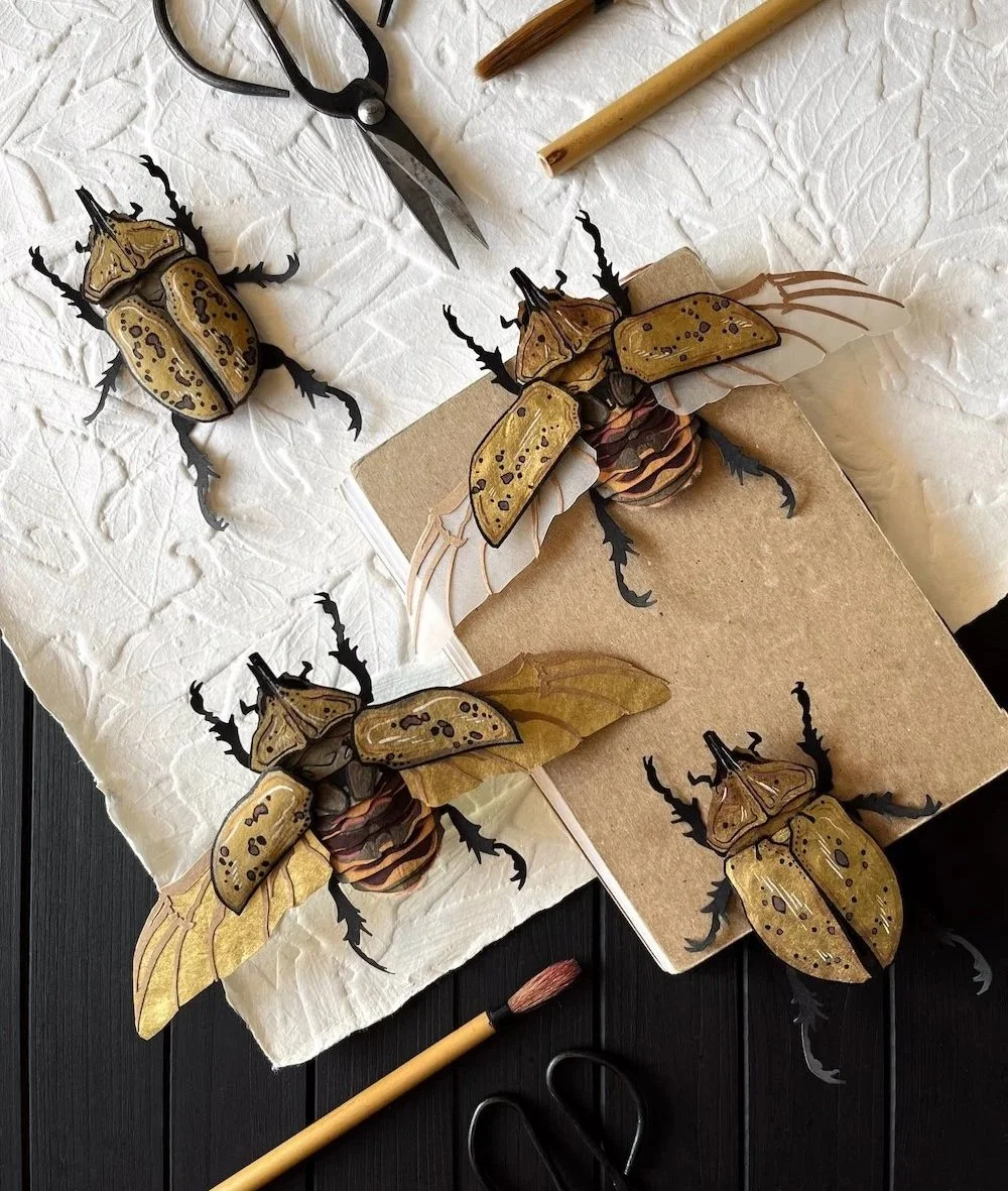 Paper Herucles beetles arranged among textured paper and a sketchbook with paintbrushes and a pair of scissors by them