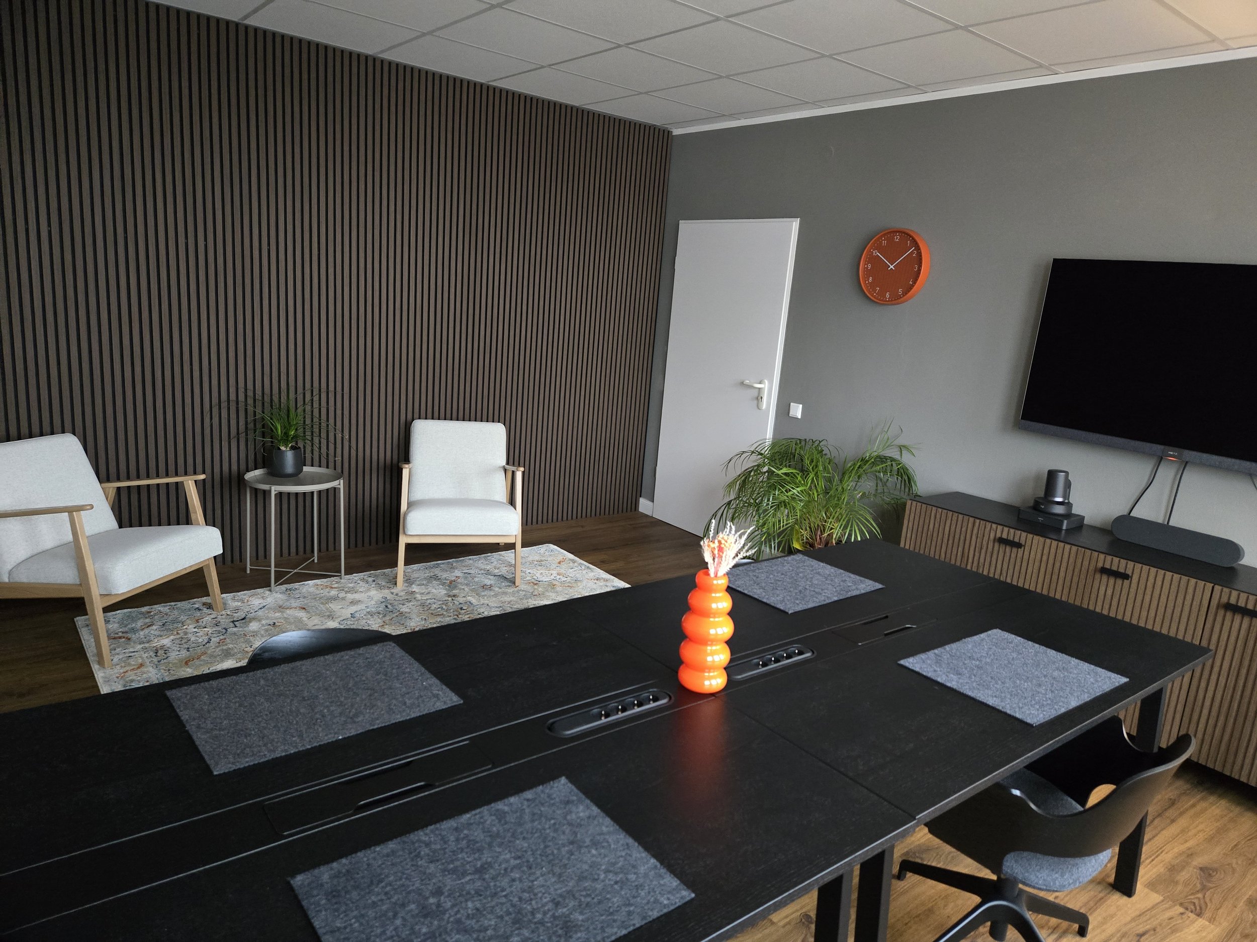Ein moderner, gut dekorierter Konferenzraum mit einem großen schwarzen Tisch, vier Sitzplätzen, einem Fernseher an der Wand, einer orangefarbenen Vase in der Mitte des Tisches, zwei Pflanzen, einem Teppich und einer Uhr an der Wand.