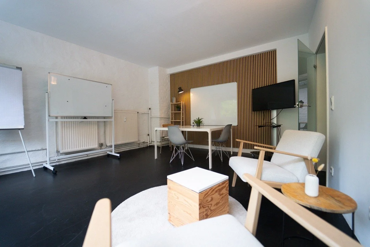 Ein modernes, minimalistisch eingerichteter Besprechungsraum mit weißen Wänden, einem schwarzen Boden, einem weißenboard, einem dekorativen Holzpaneel an der Wand, einem Fernseher, einem weißen Tisch mit Stühlen, einem weißen Sessel, einem kleinen Holz-Tablett und einem weißen Vase mit gelber Blume.