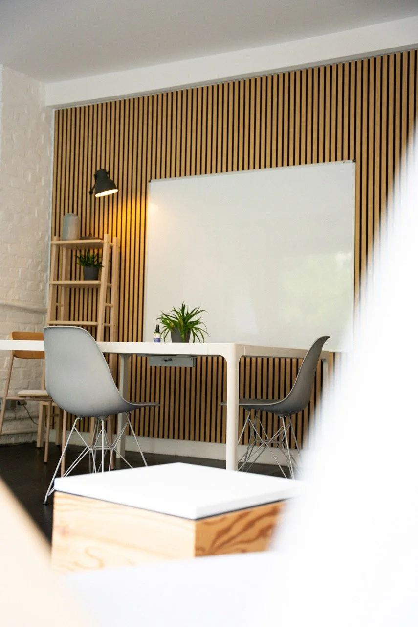 Moderner Meetingraum mit weißen Wänden, Holzpaneelwand, einem Whiteboard, zwei Stühlen, einem kleinen Tisch und Pflanzen.