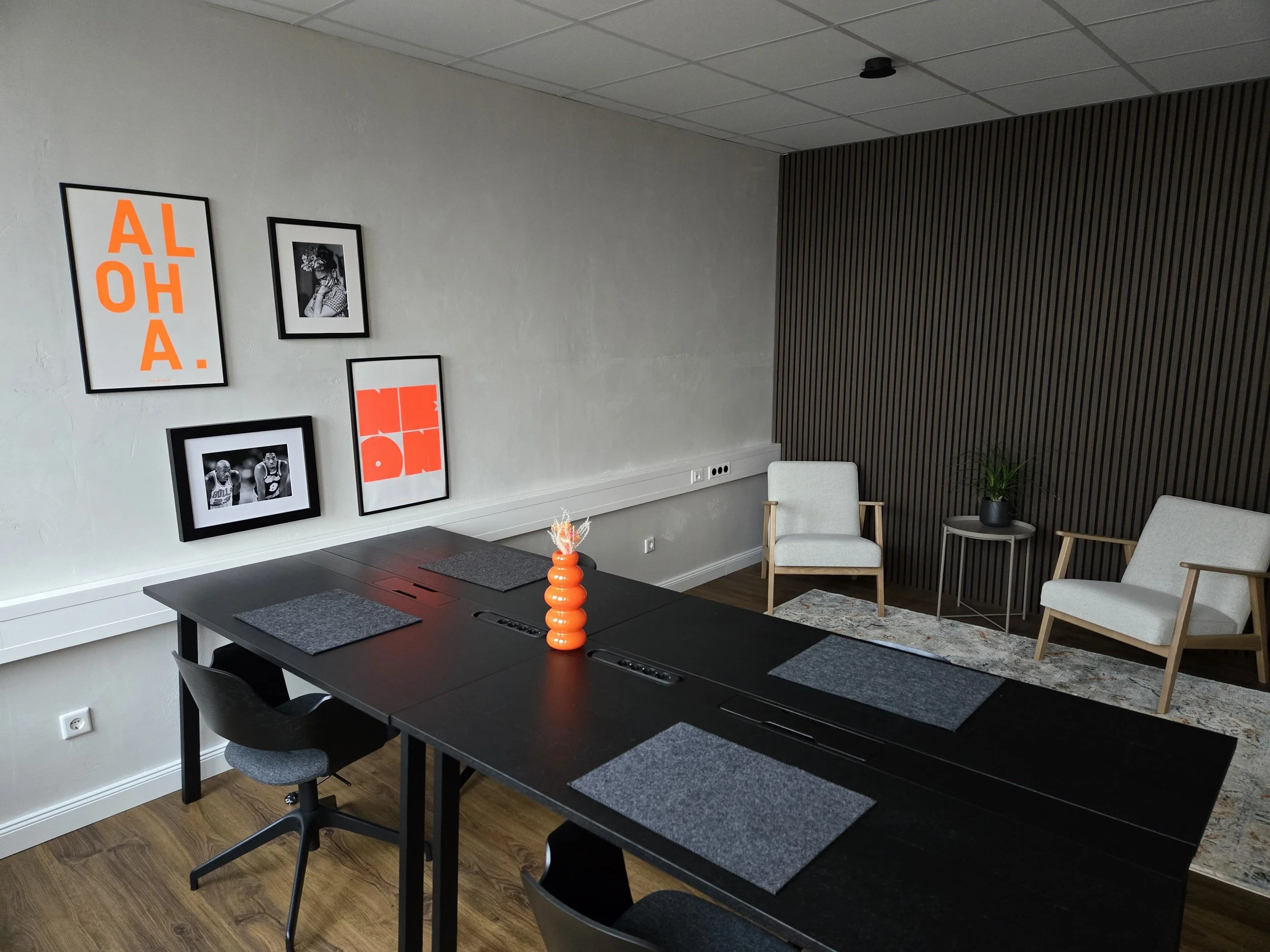 Moderner Sitzbereich mit zwei weißen Sesseln, einem Beistelltisch und einer Pflanze. Ein schwarzer Tisch mit vier Plätzen, Dekoration in einer orangen Vase, Wanddekoration mit Bilder und Spruch 'ALOHA'.