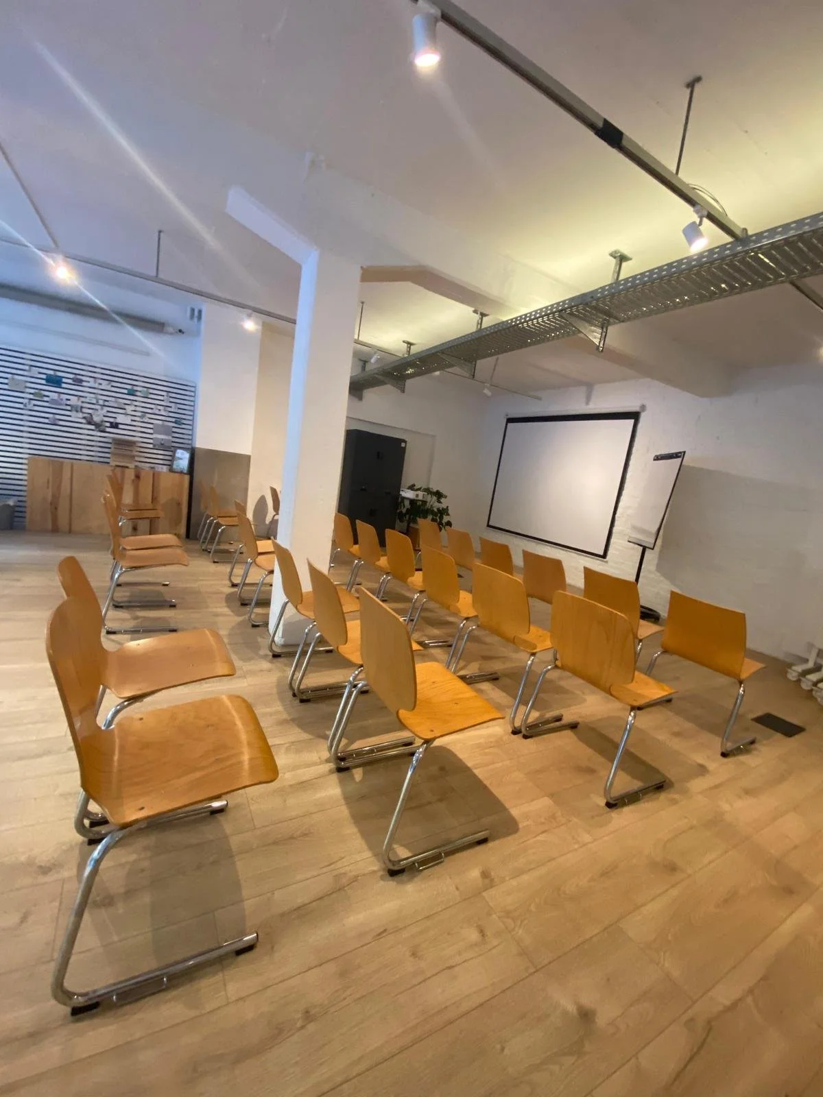 Unser vorbereiteter Eventspace, mit mehreren Reihen aus Holzstühlen mit Metallrahmen, einem großen Projektorbildschirm an der Wand und einer Whiteboard. Der Raum hat Holzfußboden und ist modern gestaltet.