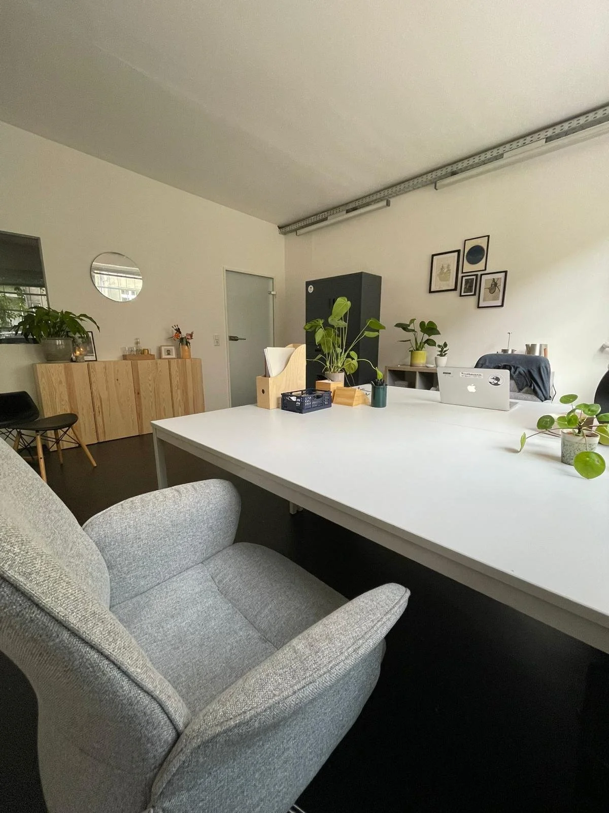 Ein moderner Büroraum mit einem großen weißen Schreibtisch, einem gemütlichen grauen Sessel, mehreren Pflanzen, einem schwarzen Schrank, Fotos an der Wand und einem Laptop.