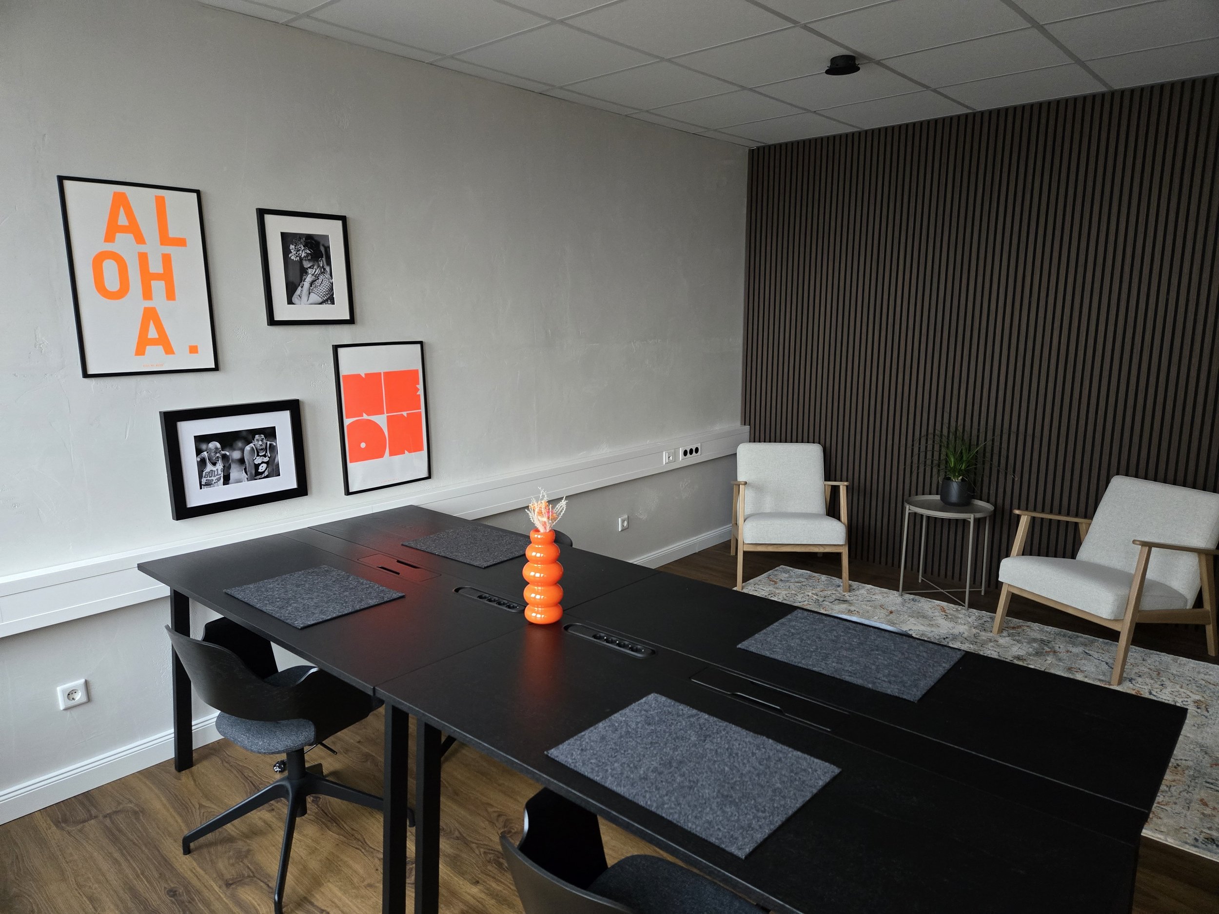 Ein moderner, minimalistisch eingerichteter Konferenzraum mit schwarzem Tisch, grauen Sitzflächen, weißen Stühlen, Wanddekoration mit Bildern und Zitaten in Schwarz, Weiß und Orange, einer Pflanze auf einem kleinen Tisch und einem modernen, dunklen Holz-Wandpaneel.