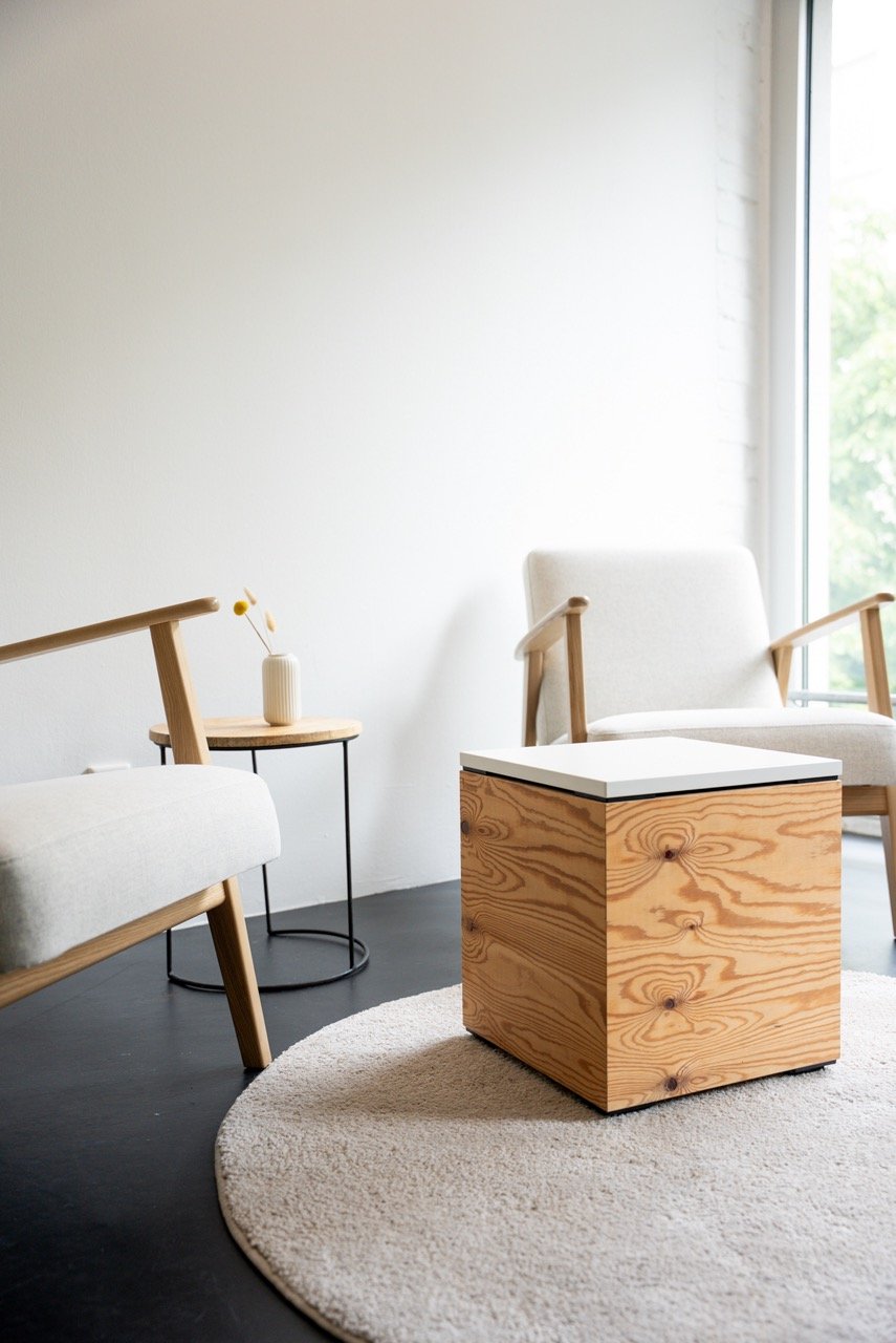 Ein moderner Meeting- und Coachingraum mit zwei gemütlichen Stühlen, einem kleinen Beistelltisch mit Vase und einer wooden storage box, alles auf einem hellen, runden Teppich, mit viel natürlichem Licht durch großes Fenster.