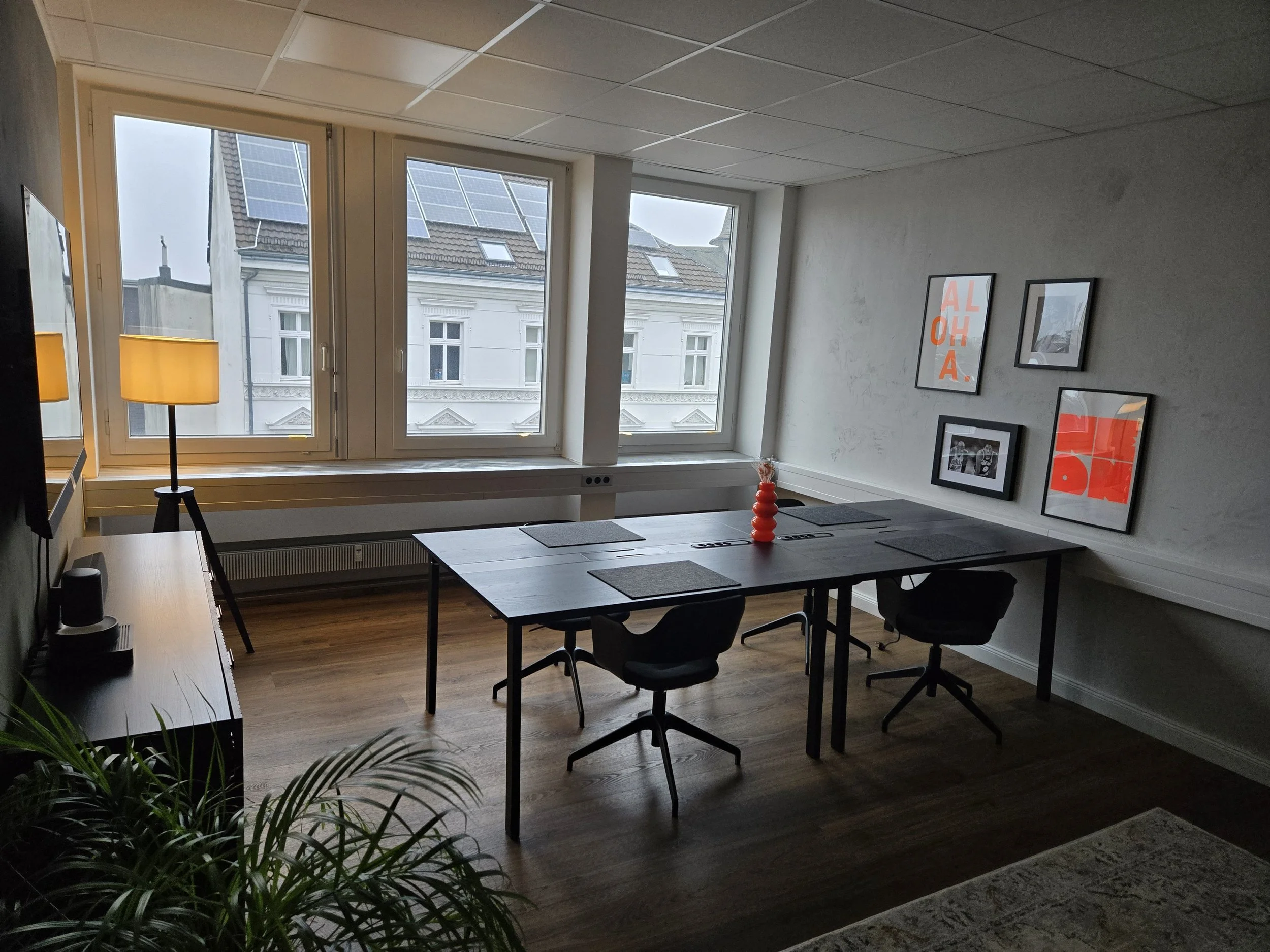 Ein modern eingerichteter Konferenzraum mit einem rechteckigen Tisch, schwarzen Stühlen und Wandbildern, große Fenster mit Blick auf alte Gebäude, ein Stehlampe und ein Fernseher