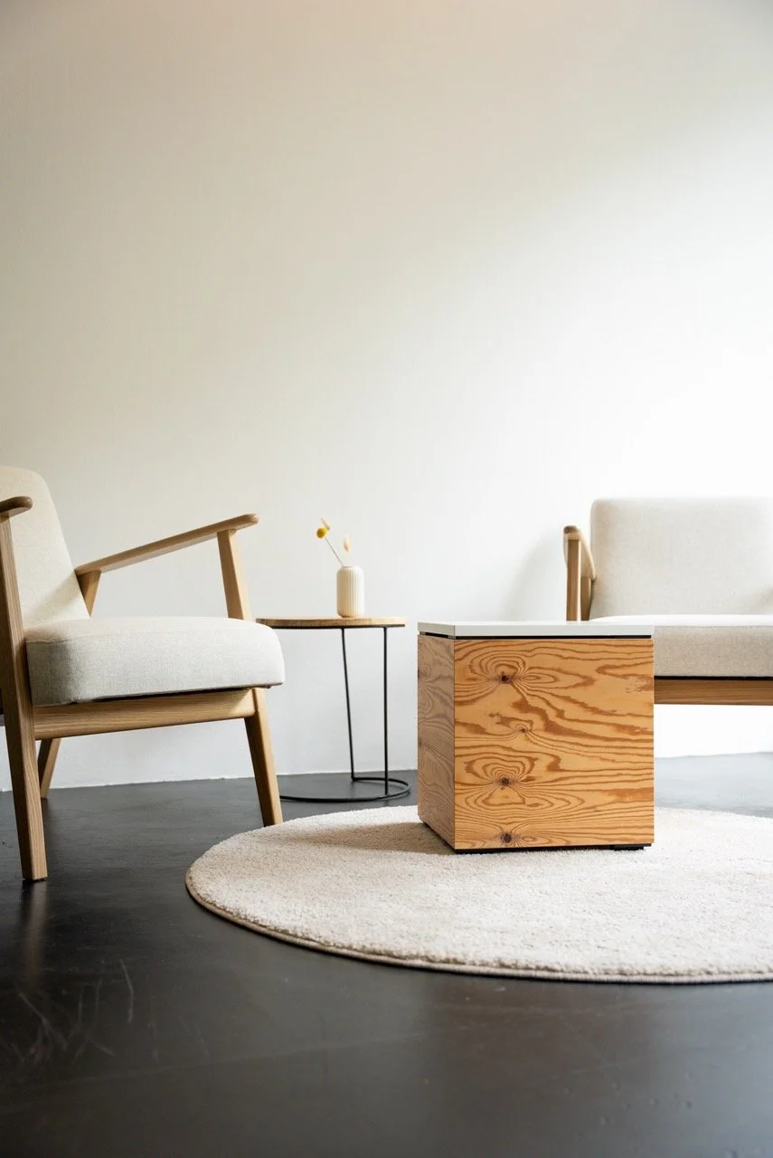 Ein minimalistisches Wohnzimmer mit holzfarbenen Möbeln, einem Sessel, einem kleinen Beistelltisch mit einer Vase, einem quadratischen Holztablett, einem weißen runden Teppich auf dunklem Boden und einer hellen Wand im Hintergrund.