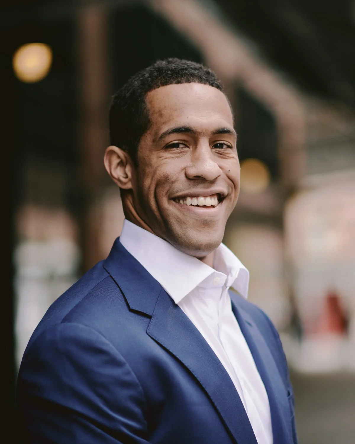 A smiling man in a navy blue suit and white shirt standing outdoors, blurred background with warm lights.