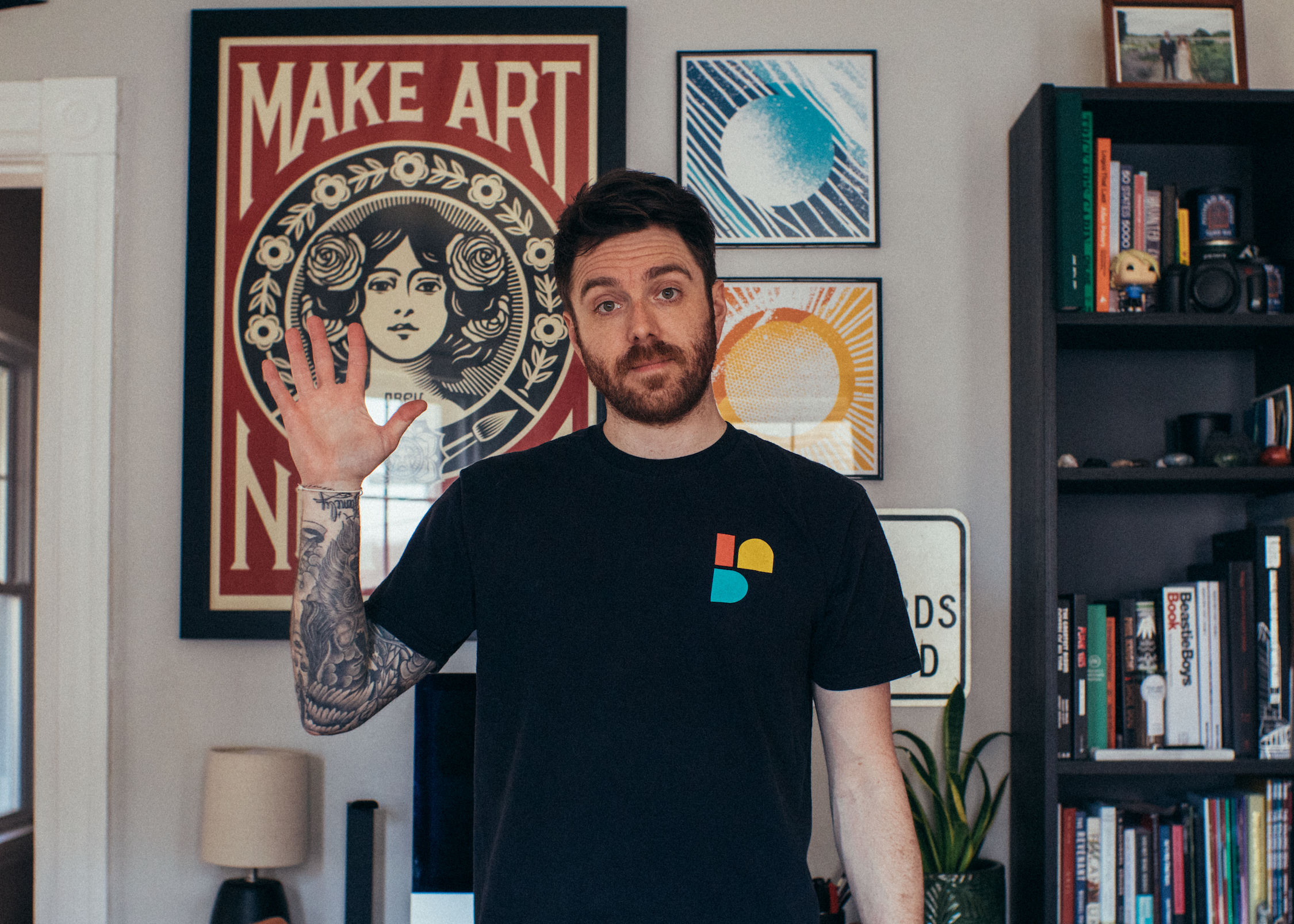 A man in a black t-shirt with a colorful logo is standing indoors, waving at the camera. Behind him are framed artwork, including a poster with a woman's face and the words "MAKE ART," and other geometric designs. The background also features a bookshelf filled with books and decorative items, and a lamp.