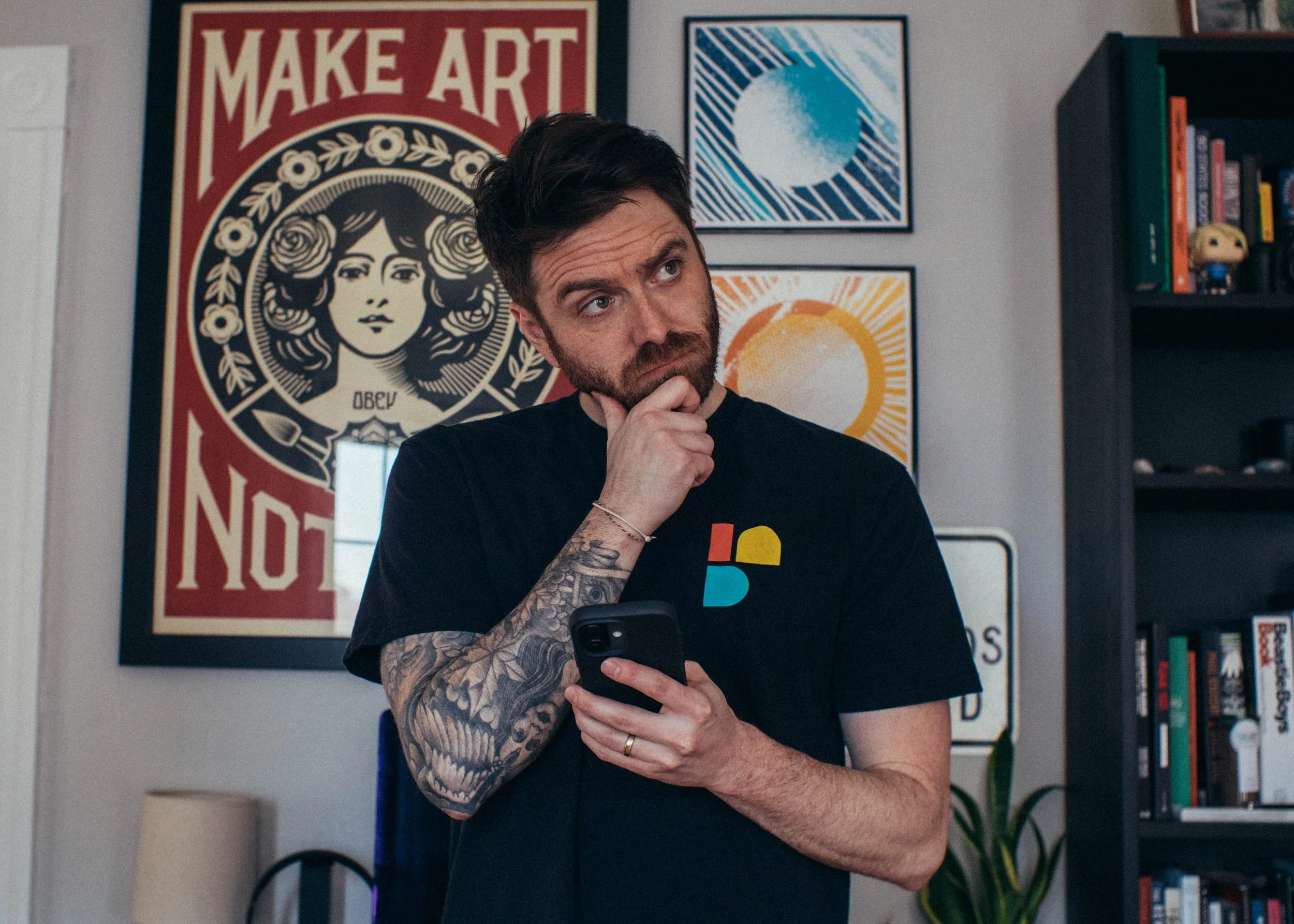 A man with dark hair and tattoos on his right arm, holding a smartphone and looking to the side, standing in a room with colorful artwork and a bookshelf in the background.
