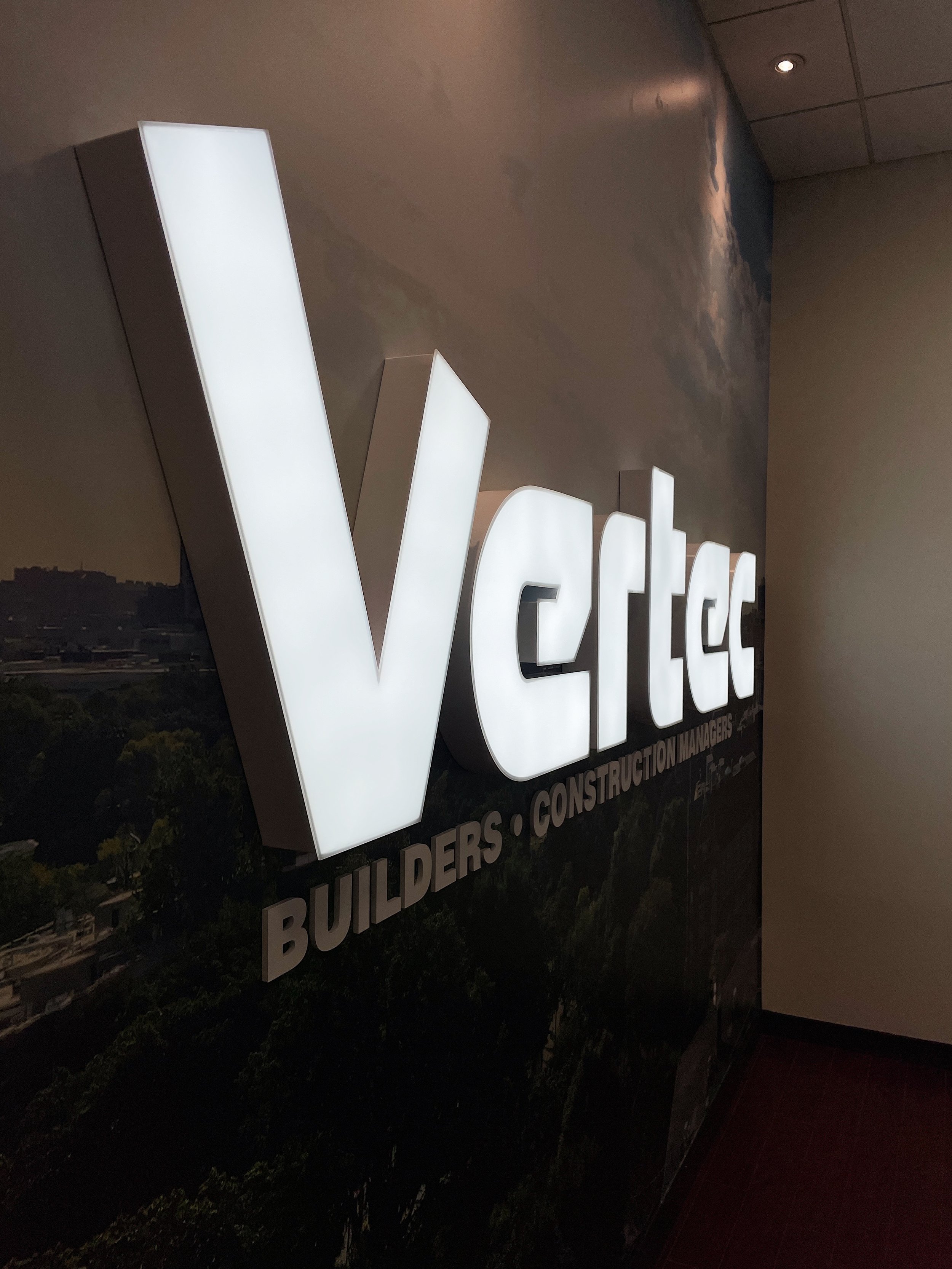 Illuminated sign for Vertec, a company specializing in builders and construction management, mounted on a wall with a scenic background.