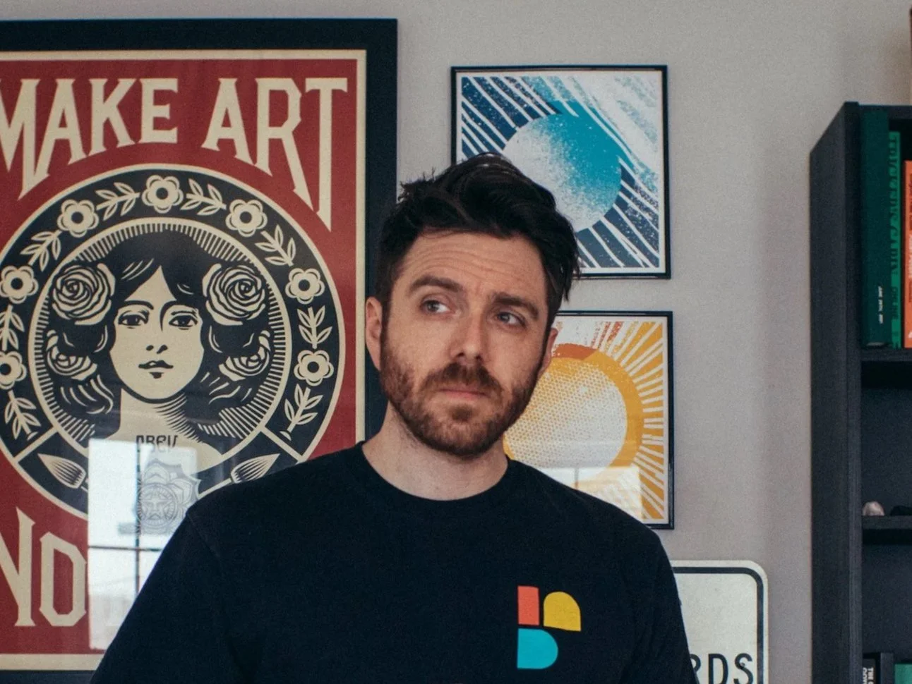 Man with dark hair and beard standing in front of wall decorated with art posters, including a large red and black poster saying "MAKE ART" and two smaller posters of stylized sun and moon.