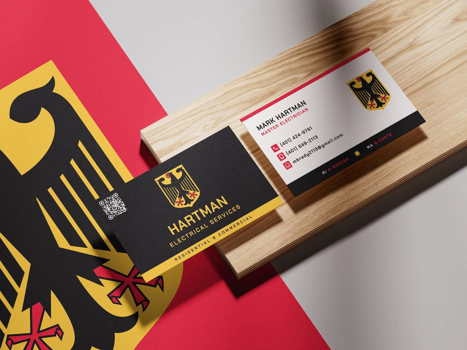 Business cards for Hartman Electrical Services, featuring a black and yellow eagle logo, contact details, and a QR code, placed on a wooden surface with a red, white, and black background.