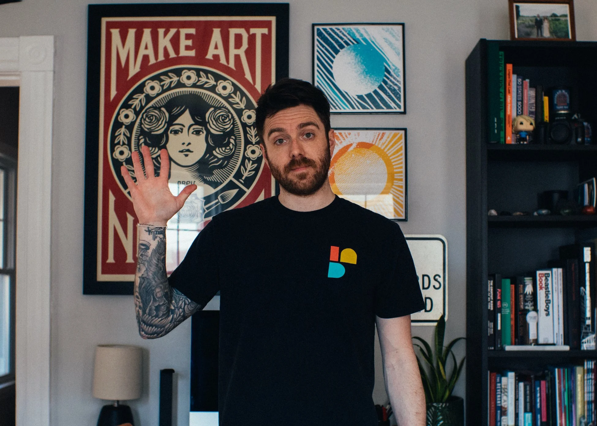 A man with dark hair and a beard waving in a living room, wearing a black t-shirt with a colorful logo, standing in front of a wall with framed artwork and a bookshelf filled with books and small collectibles.