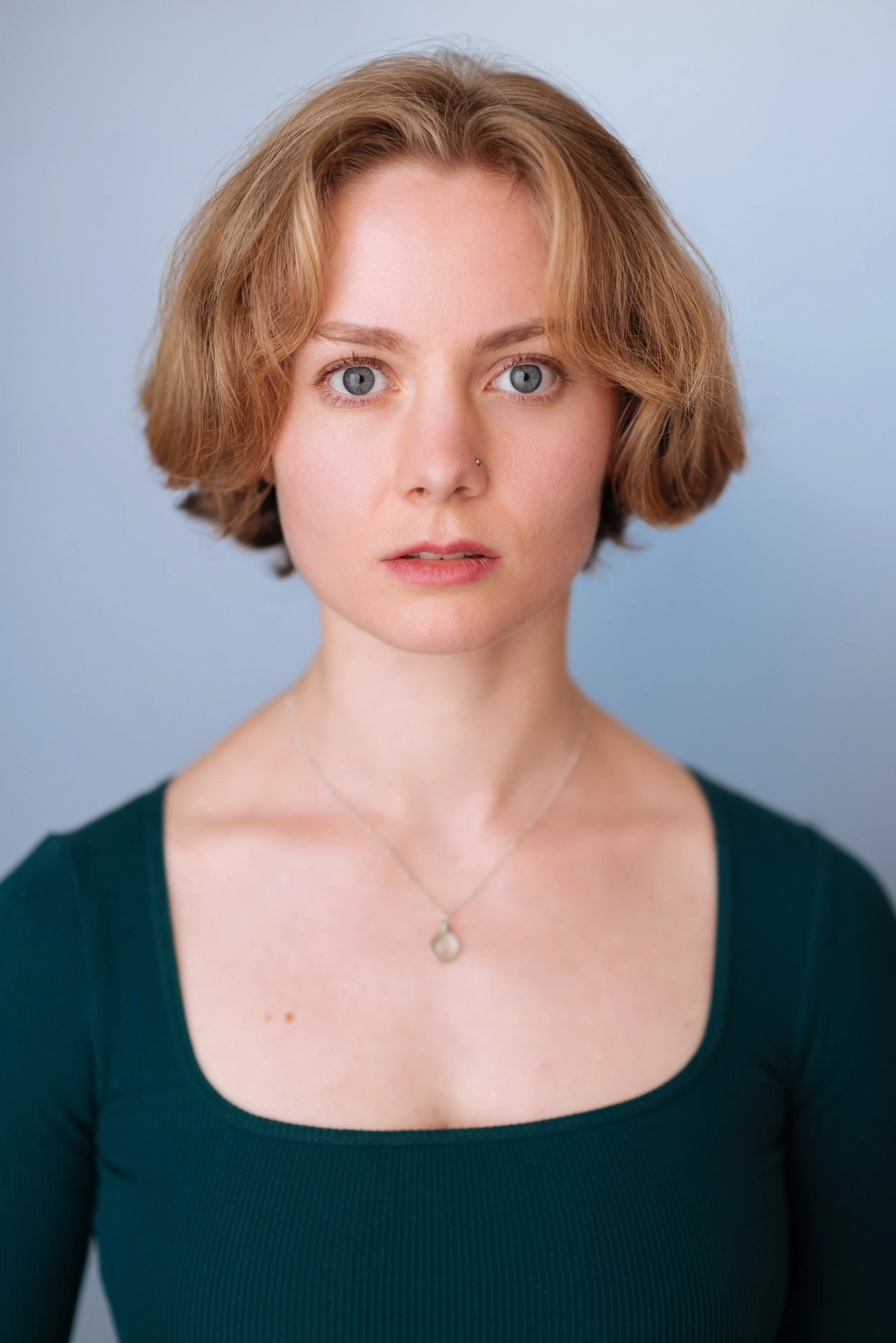 Headshot of Emily Cummings, an actress with short, wavy blonde hair, blue eyes, wearing a dark green top and a pearl necklace, against a light blue background. In the shot, she looks dramatic and vulnerable.