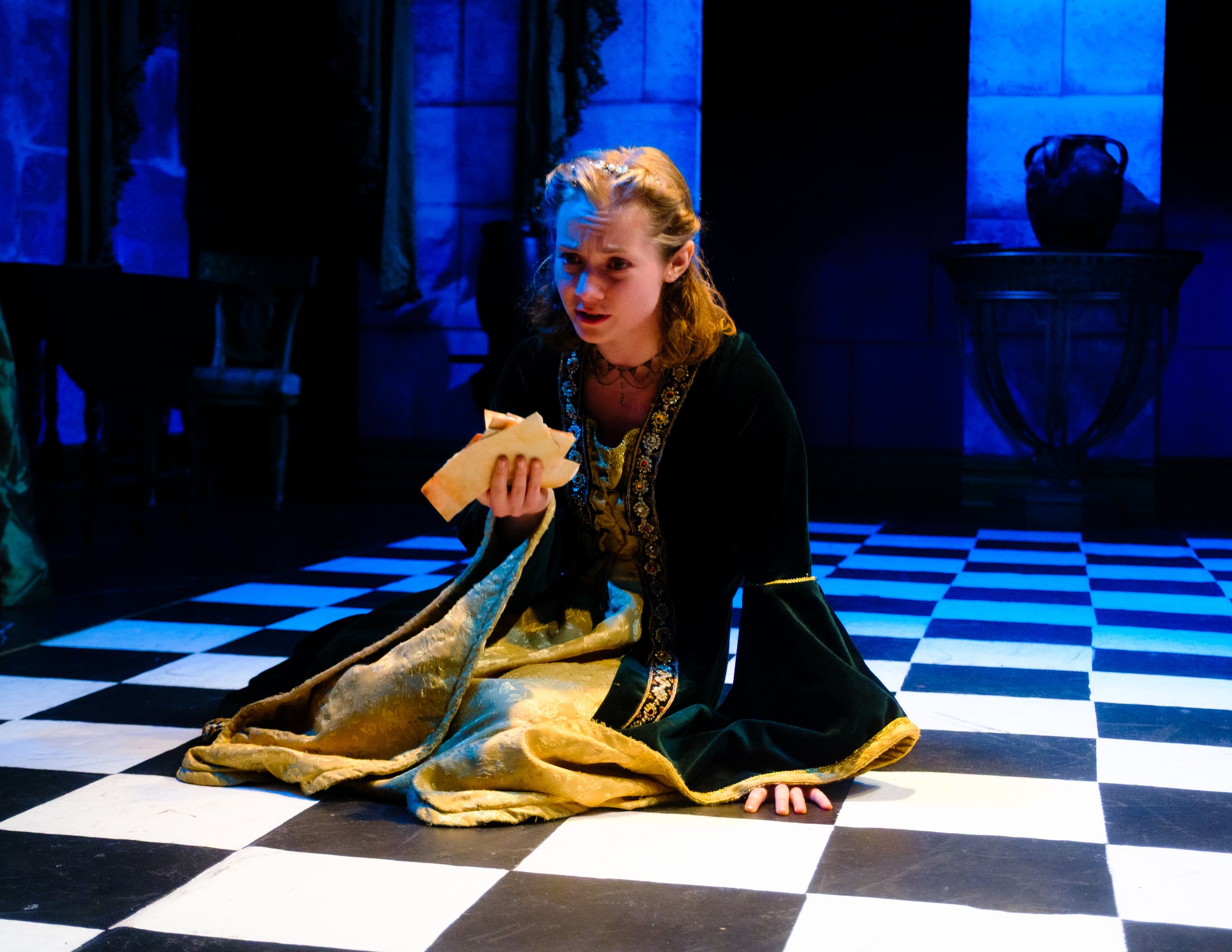 A photo of a theatrical performance in which a young actress, Emily Cummings, dressed in an ornate costume, sits on a checkered black and white floor in a scene lit with blue lighting, holding a piece of paper or a letter.