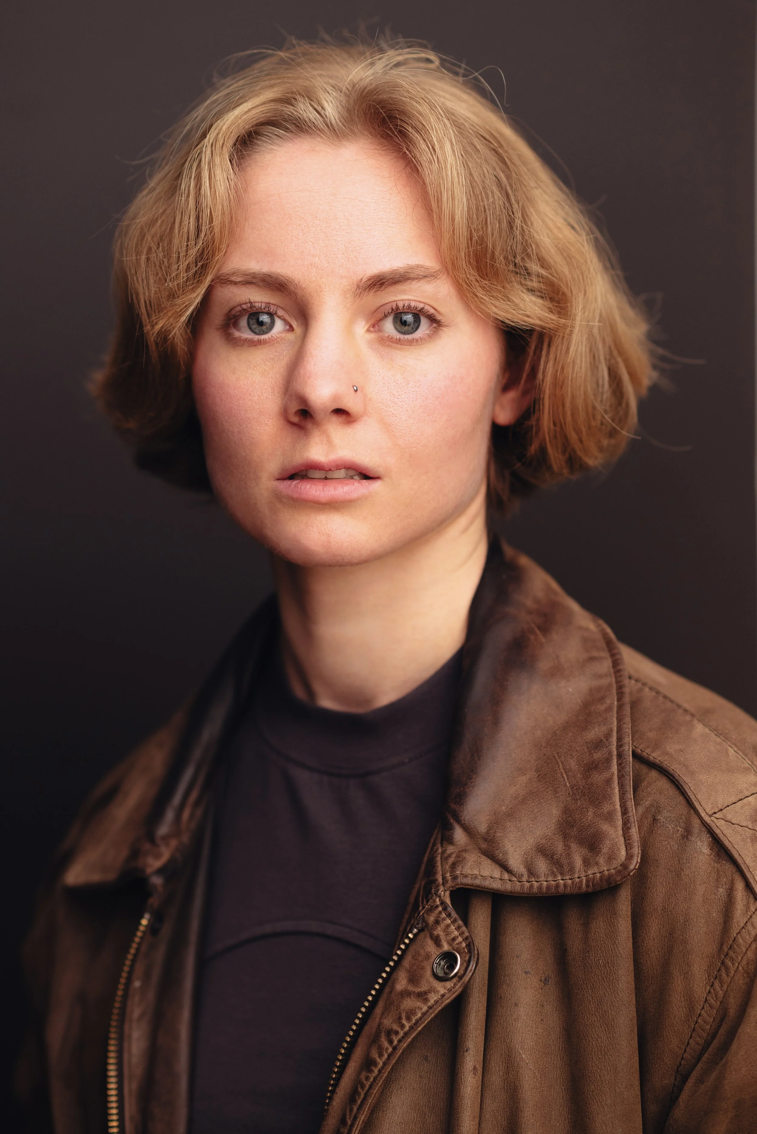 Headshot of a Emily Cummings, an actor with short blond hair, blue eyes, and a nose piercing, wearing a brown leather jacket and a black shirt against a dark background. In the shot she looks tough, pensive, rugged, and determined.