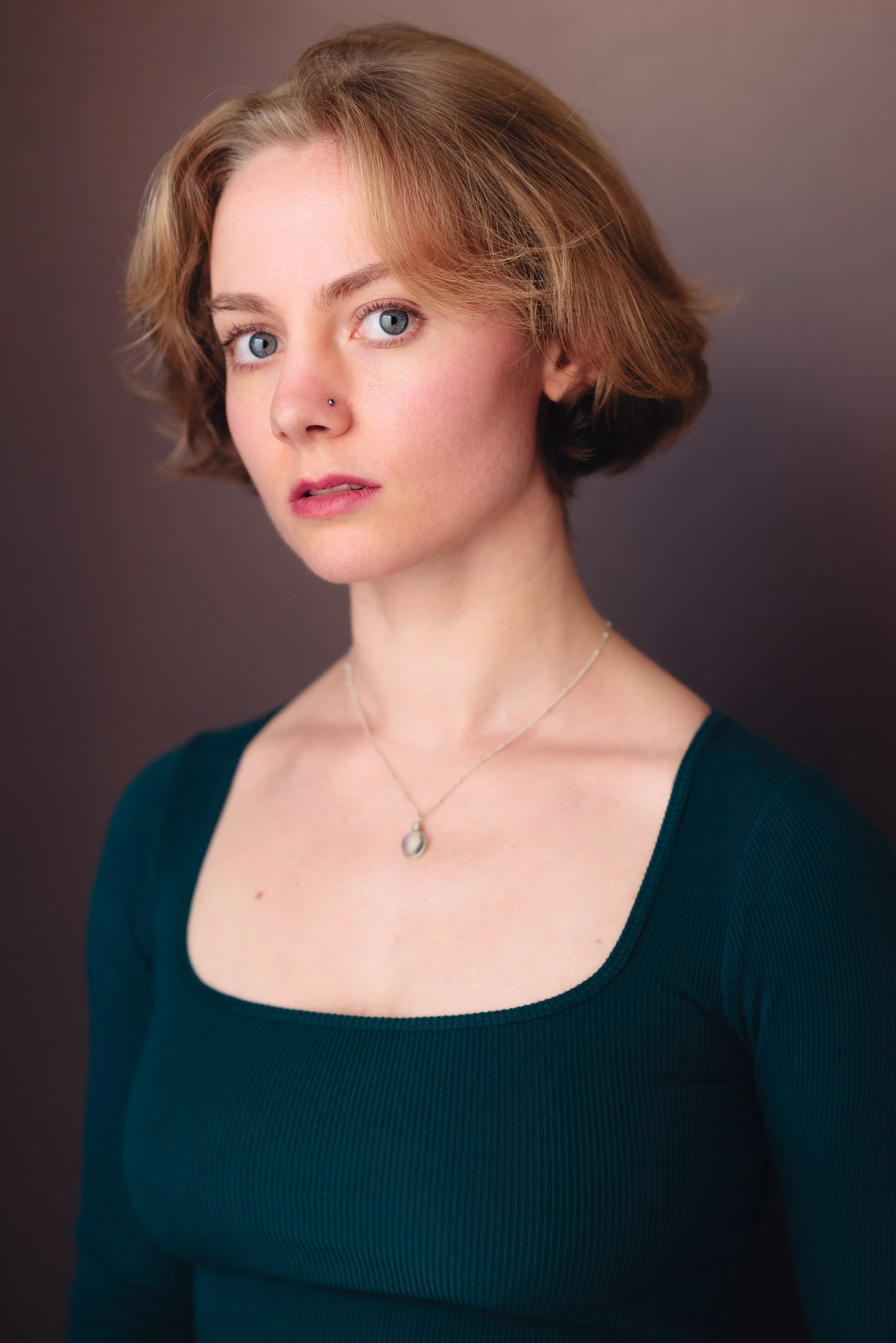 Headshot of Emily Cummings, an actress woman with short, wavy  blond hair and blue eyes, wearing a green top and a silver necklace, with a gray background. She looks pretty and curious, like a romantic lead.