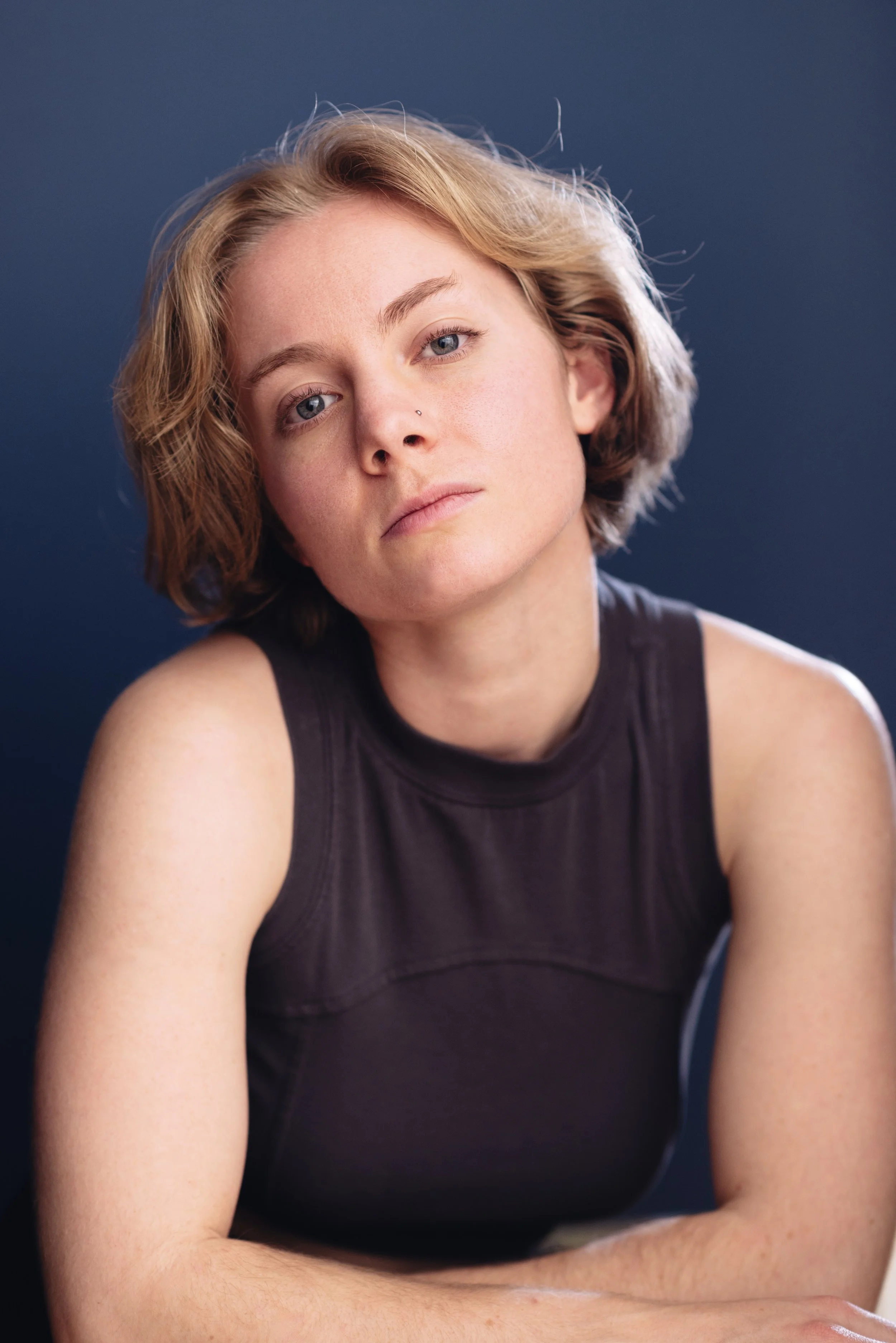 Headshot of a young actress, Emily Cummings with short, wavy blonde hair, blue eyes, and a nose piercing, wearing a sleeveless black top, against a dark blue background. In the shot, she looks tough, athletic, tomboyish, and confident.
