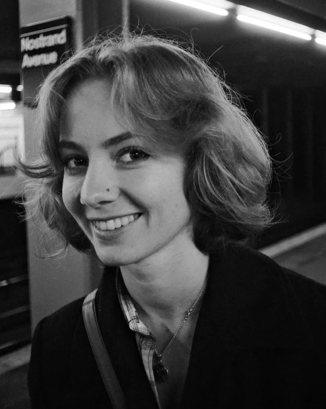 A black-and-white photo of a young woman, actress Emily Cummings, with wavy hair smiling at the camera inside a train station, with a sign in the background that reads 'Nostrand Avenue'.