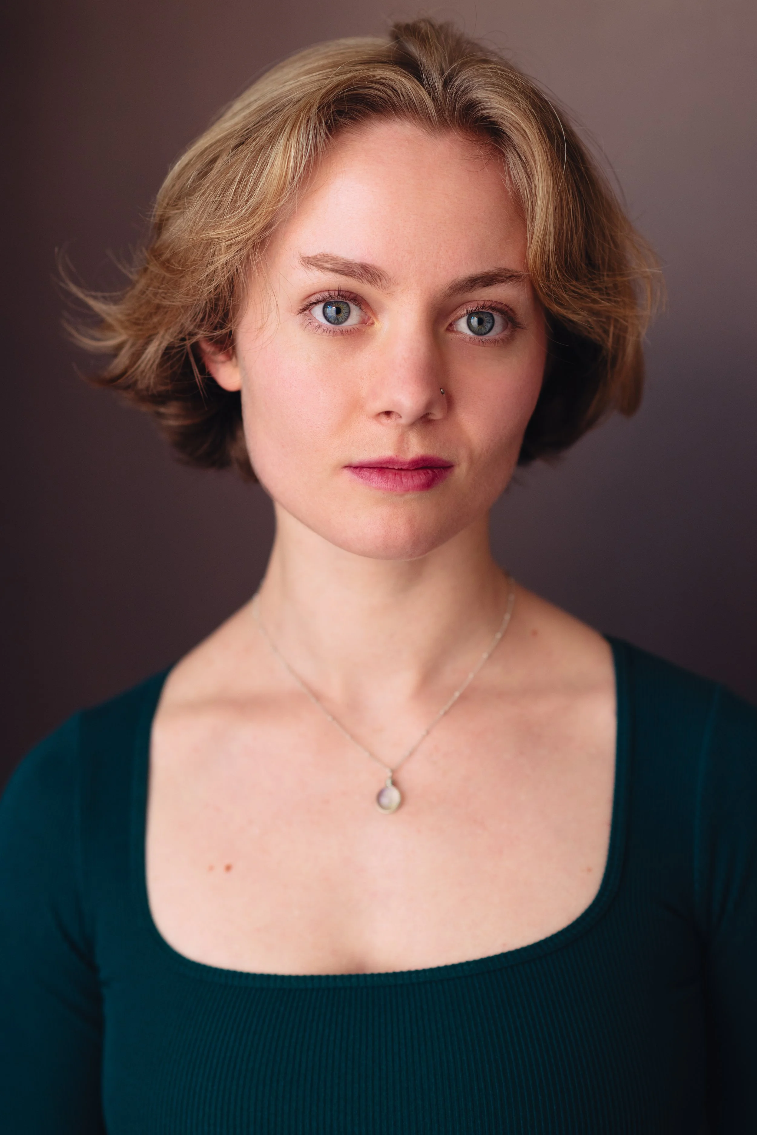 Headshot of a young actress, Emily Cummings, with short blonde hair and blue eyes, wearing a dark teal top and a pearl necklace, looking directly at the camera with a neutral expression. She looks gentle, kind, and loving.