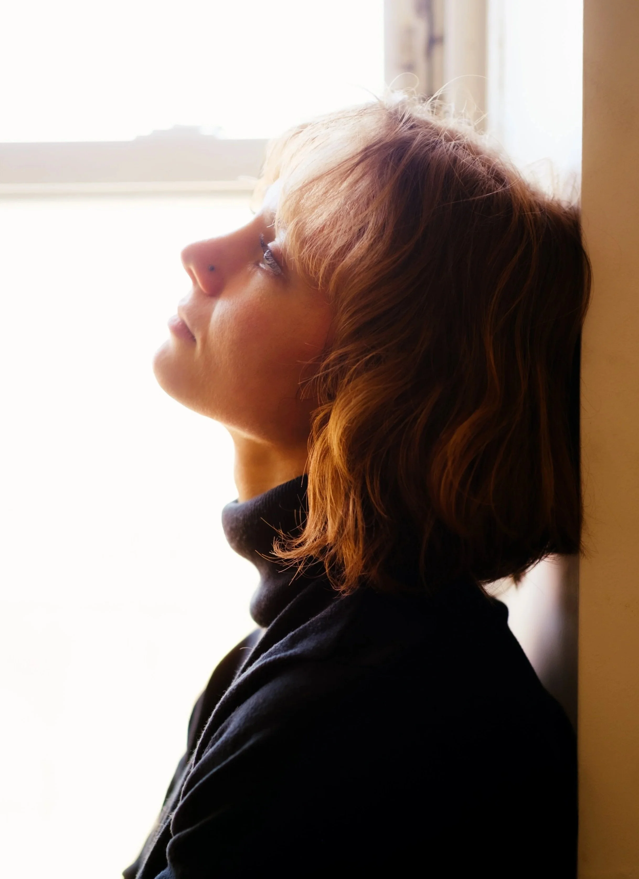 Side profile of a Emily Cummings, an actress with blond, wavy hair, wearing a black top, looking toward a bright window with sunlight streaming in.