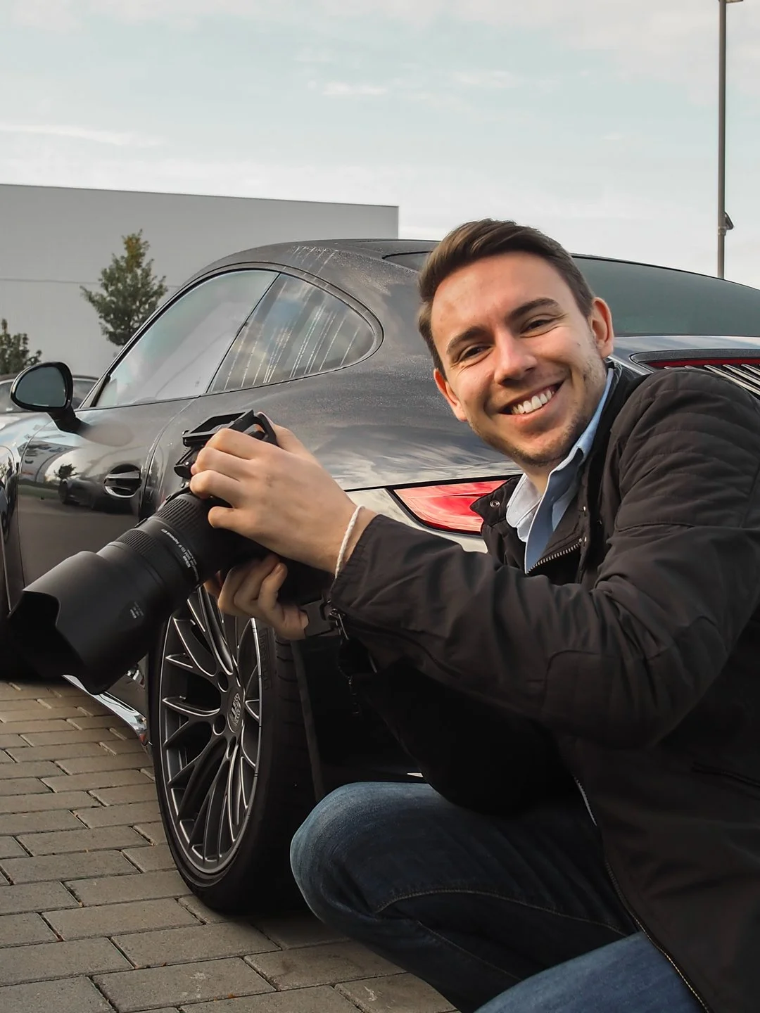 Junge Mann mit Kamera neben schwarzem Auto, lächelnd, draußen, in der Nähe eines Gebäudes.