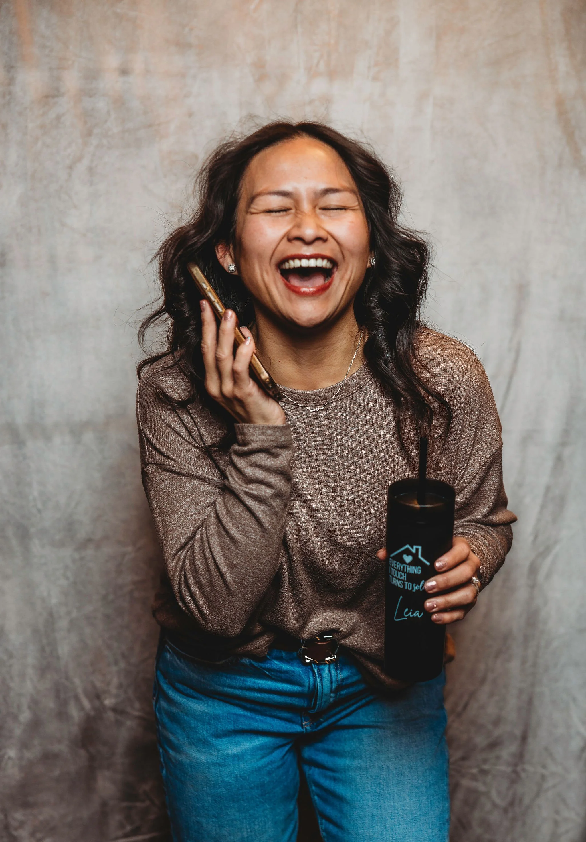 A woman with long dark hair, smiling and laughing, holding a cup with a straw. She is wearing a black jacket and is standing in front of a mirror in a store, with other items and a shopping cart nearby.