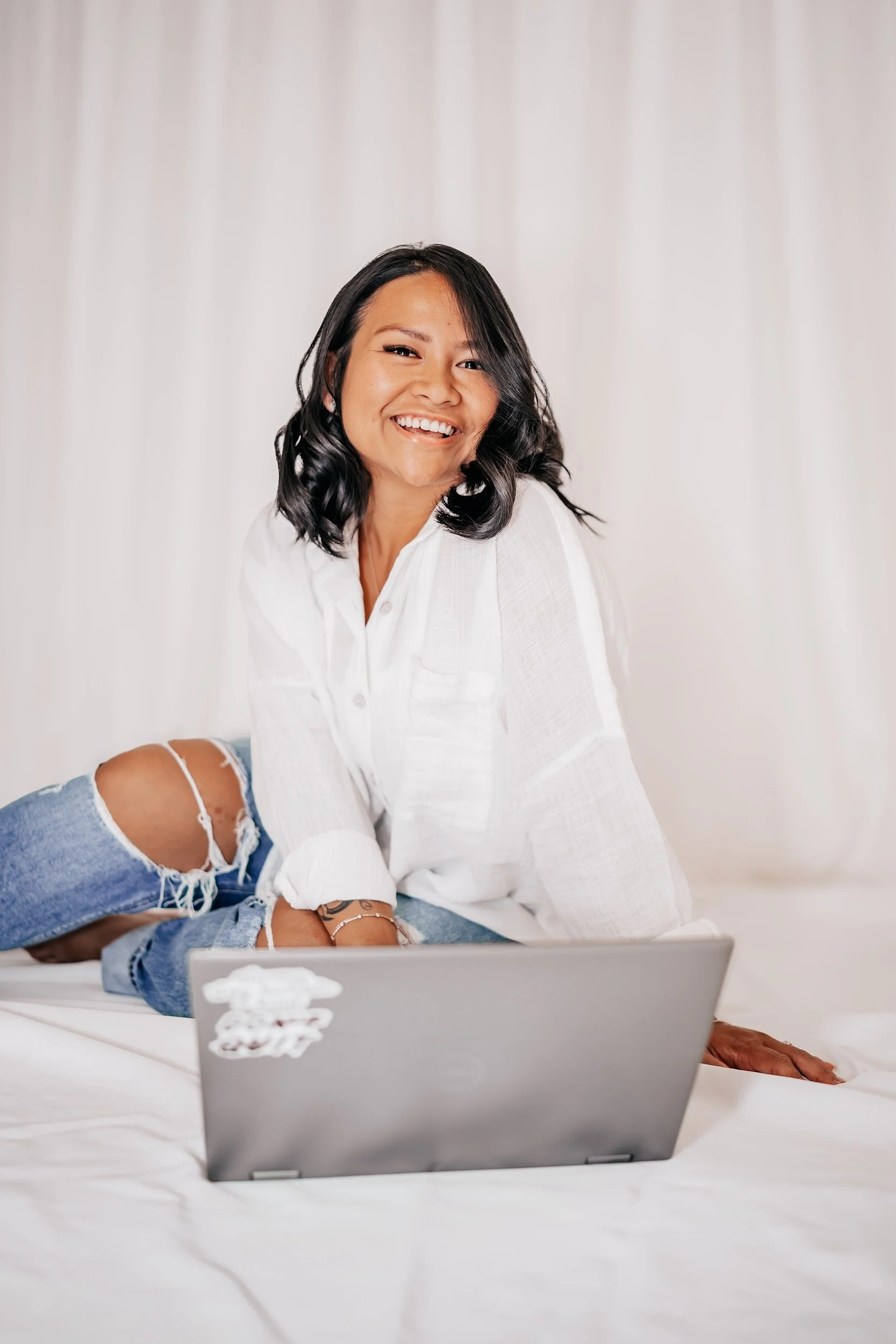 A woman with shoulder-length black hair, wearing a white shirt, is smiling and sitting on a bed with a laptop in front of her, against a white curtain background.