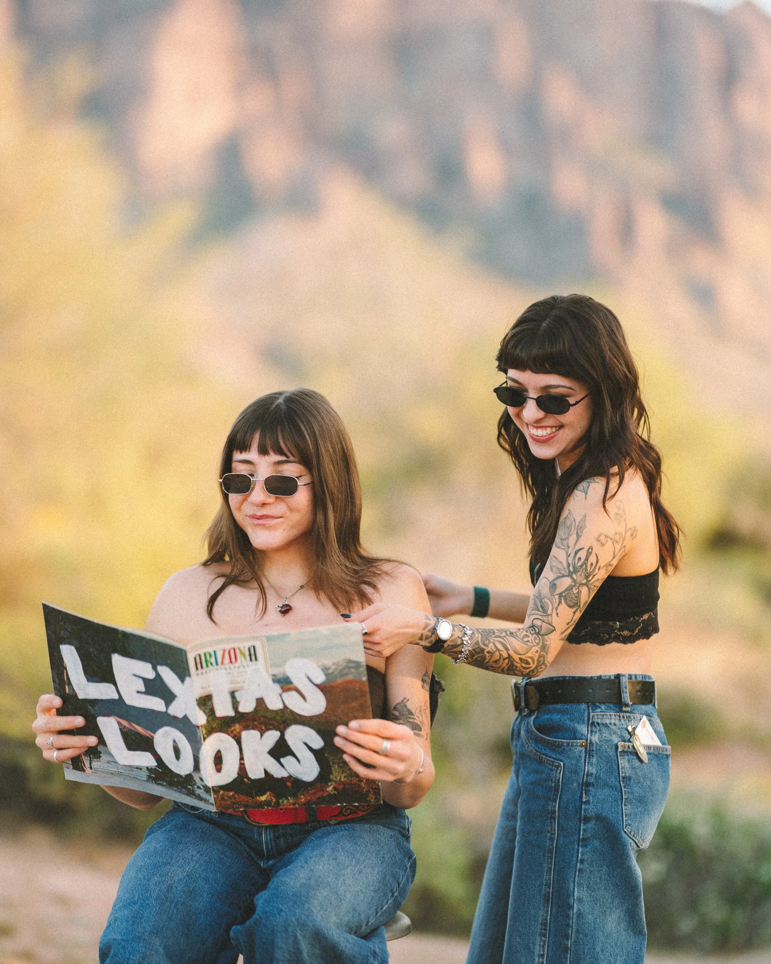 Two women with tattoos and sunglasses, one sitting and reading a map while the other stands and points to it, in outdoor scenery with mountains in the background.