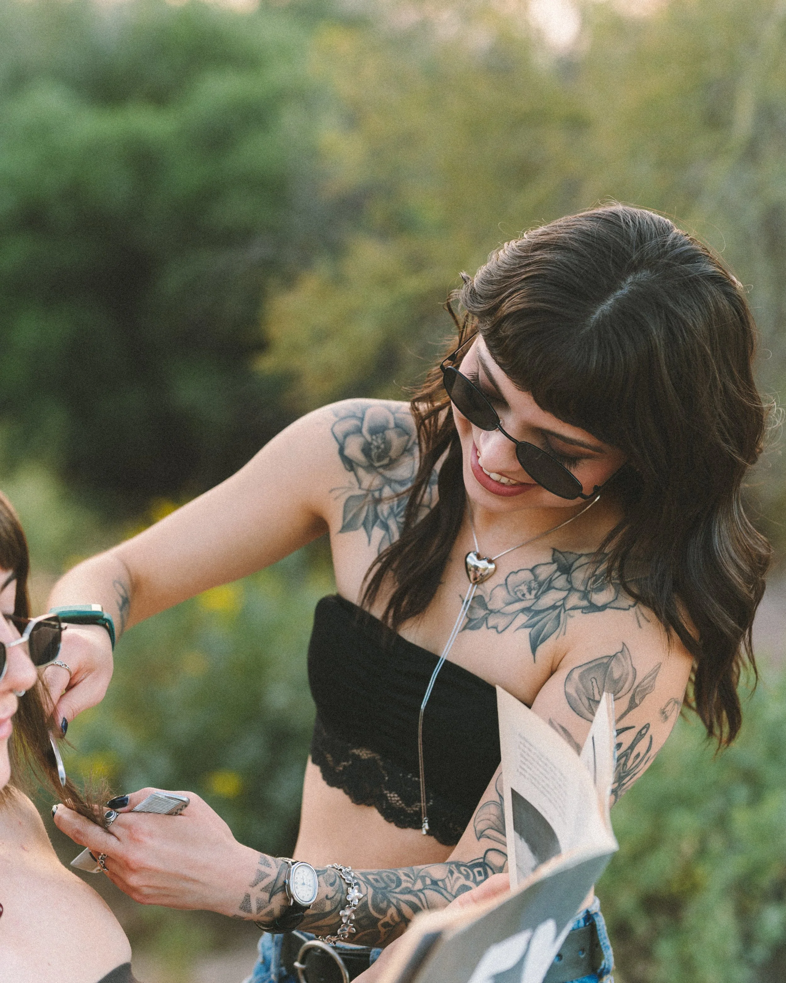 A young woman with dark hair, sunglasses, and tattoos on her arms and chest, wearing a black tube top, smiling while looking at a phone outdoors.