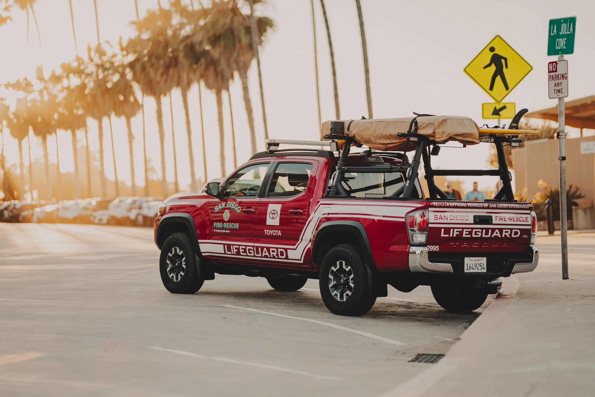 A red Toyota Tacoma pickup truck with San Diego Fire-Rescue and Lifeguard markings, parked along a street with palm trees in the background, and traffic signs including a pedestrian crossing and no parking signs visible.
