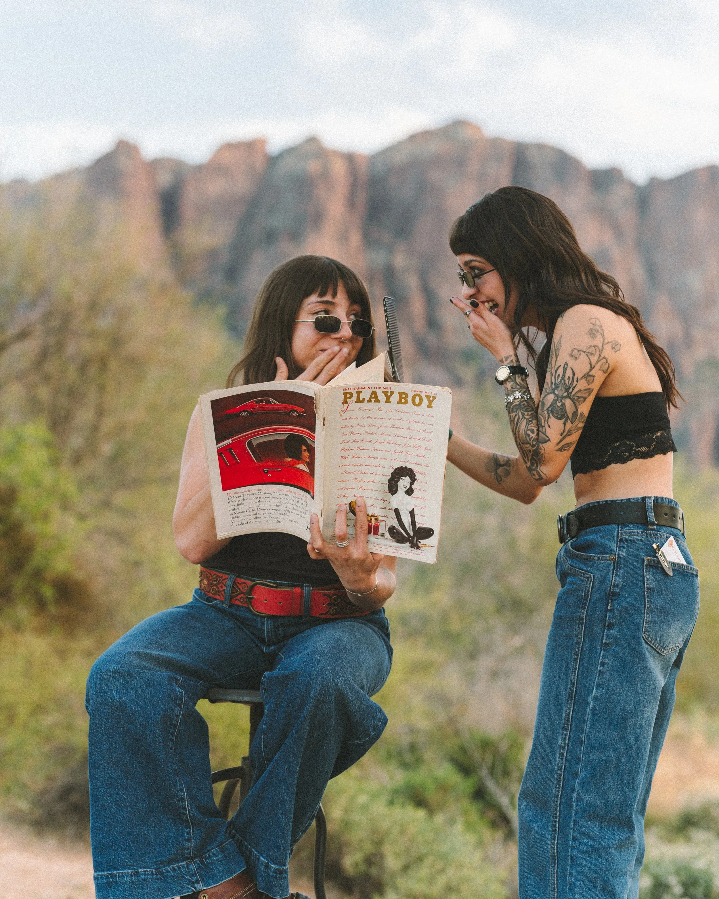 Two women outdoors, one sitting on a chair and holding a Playboy magazine, the other standing and covering her mouth as if sharing a secret or laughing, with a scenic mountain background.