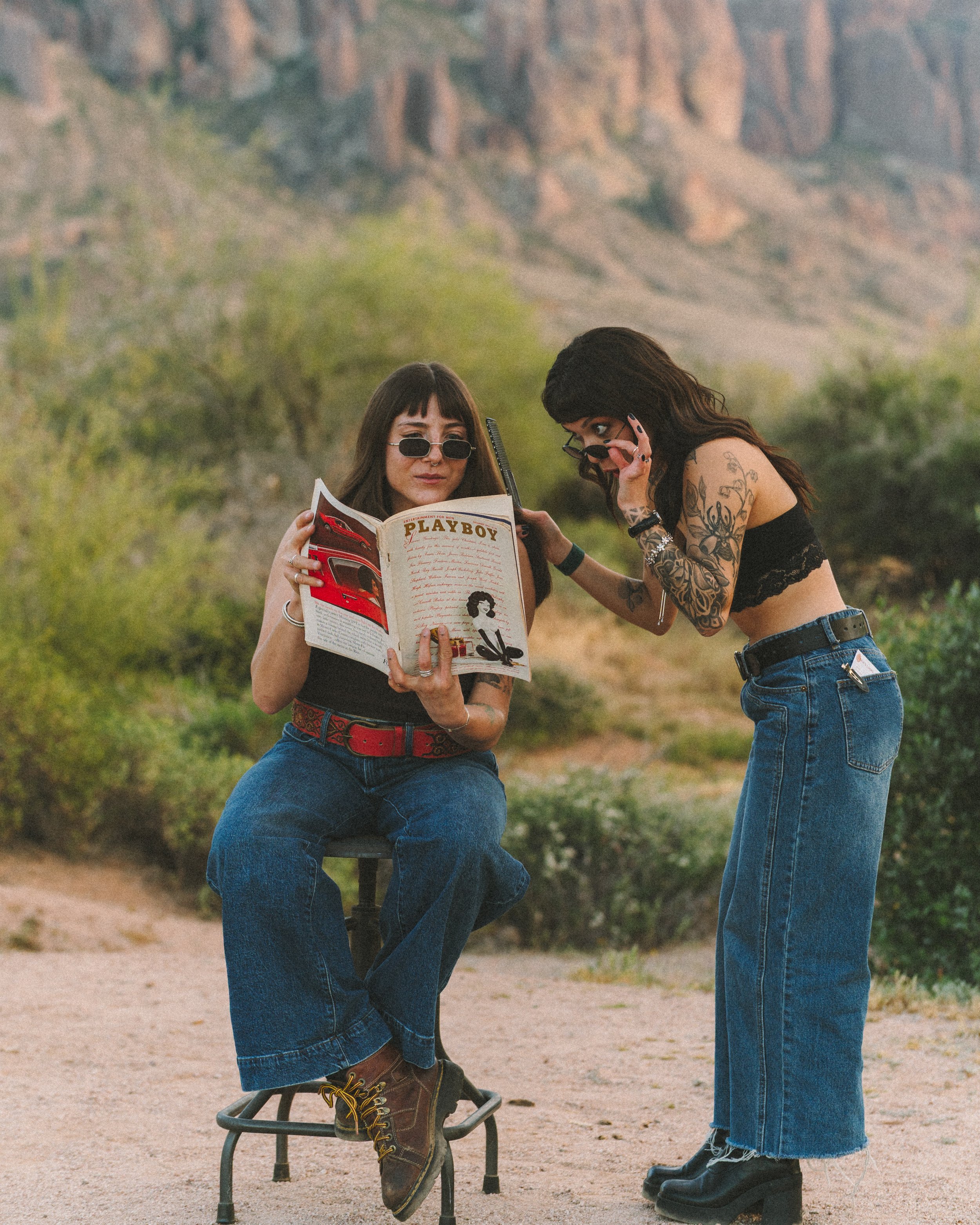 Two women with dark hair and tattoos, one seated on a stool reading a Playboy magazine, the other leaning in to look at the magazine, with a desert landscape and rocky cliffs in the background.