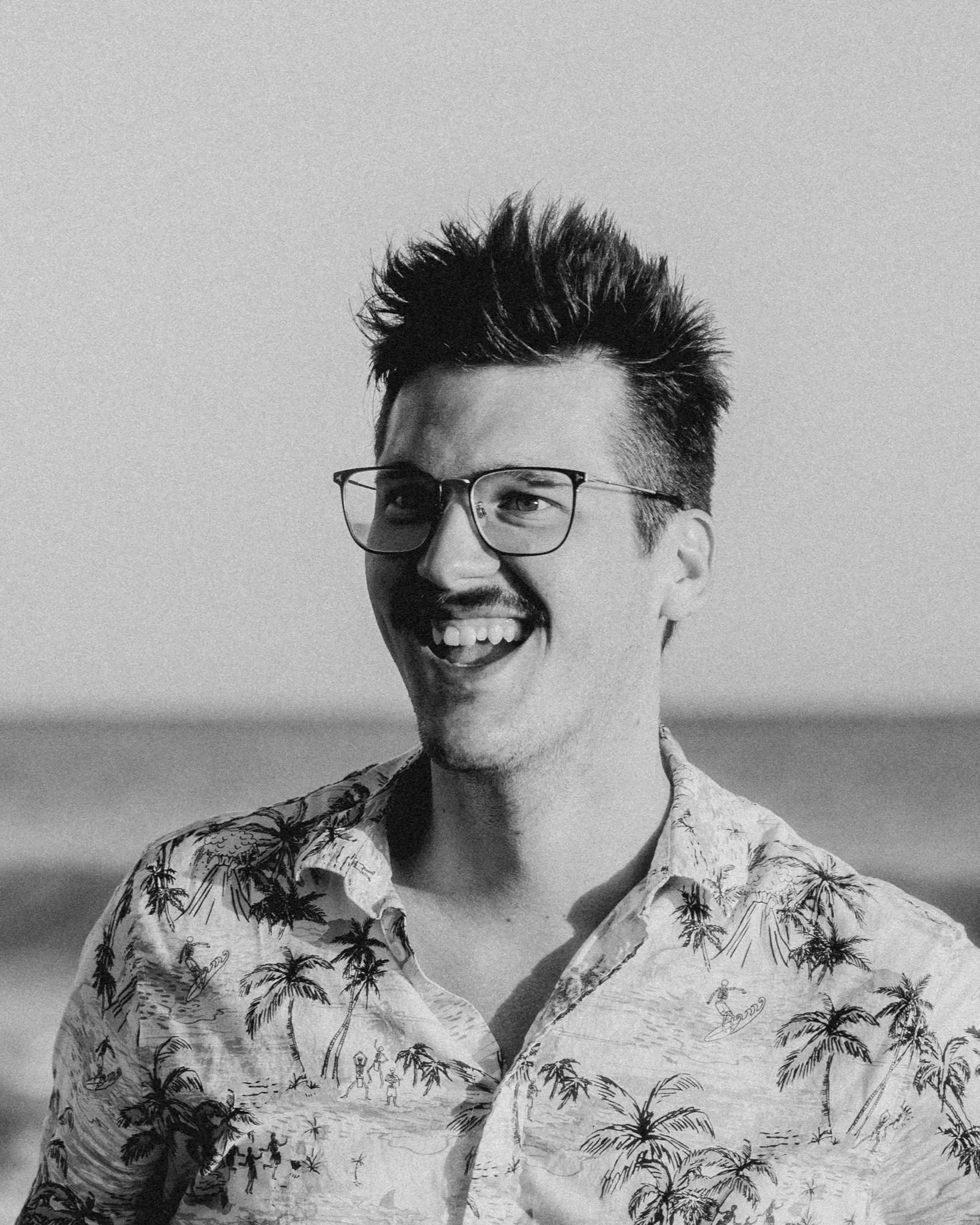 Black and white photo of a smiling man with glasses, wearing a tropical-themed shirt, outdoors with a blurry background.