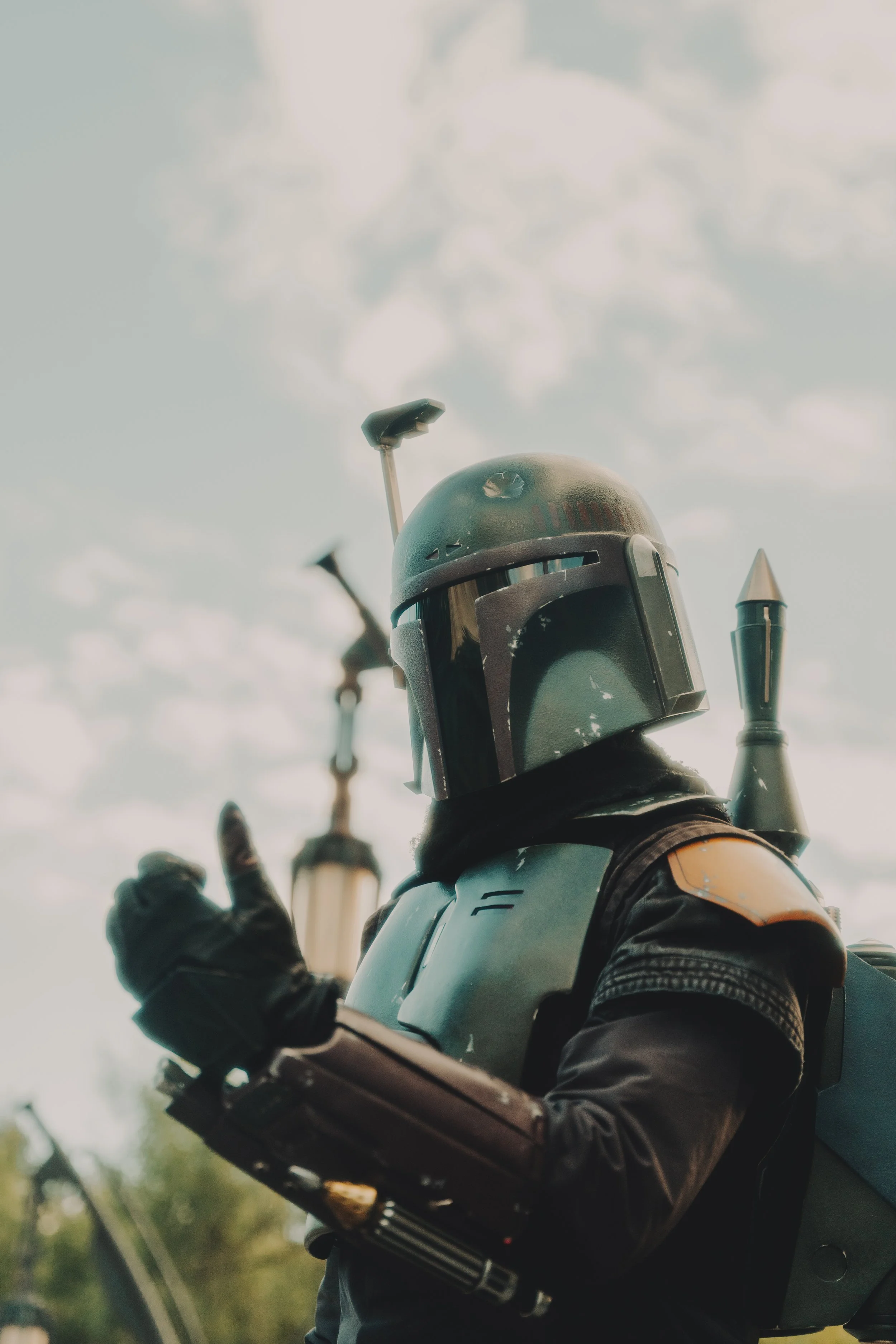 A person dressed as Boba Fett from Star Wars, wearing a helmet and armor, making a peace sign with their left hand, outdoors with a cloudy sky in the background.