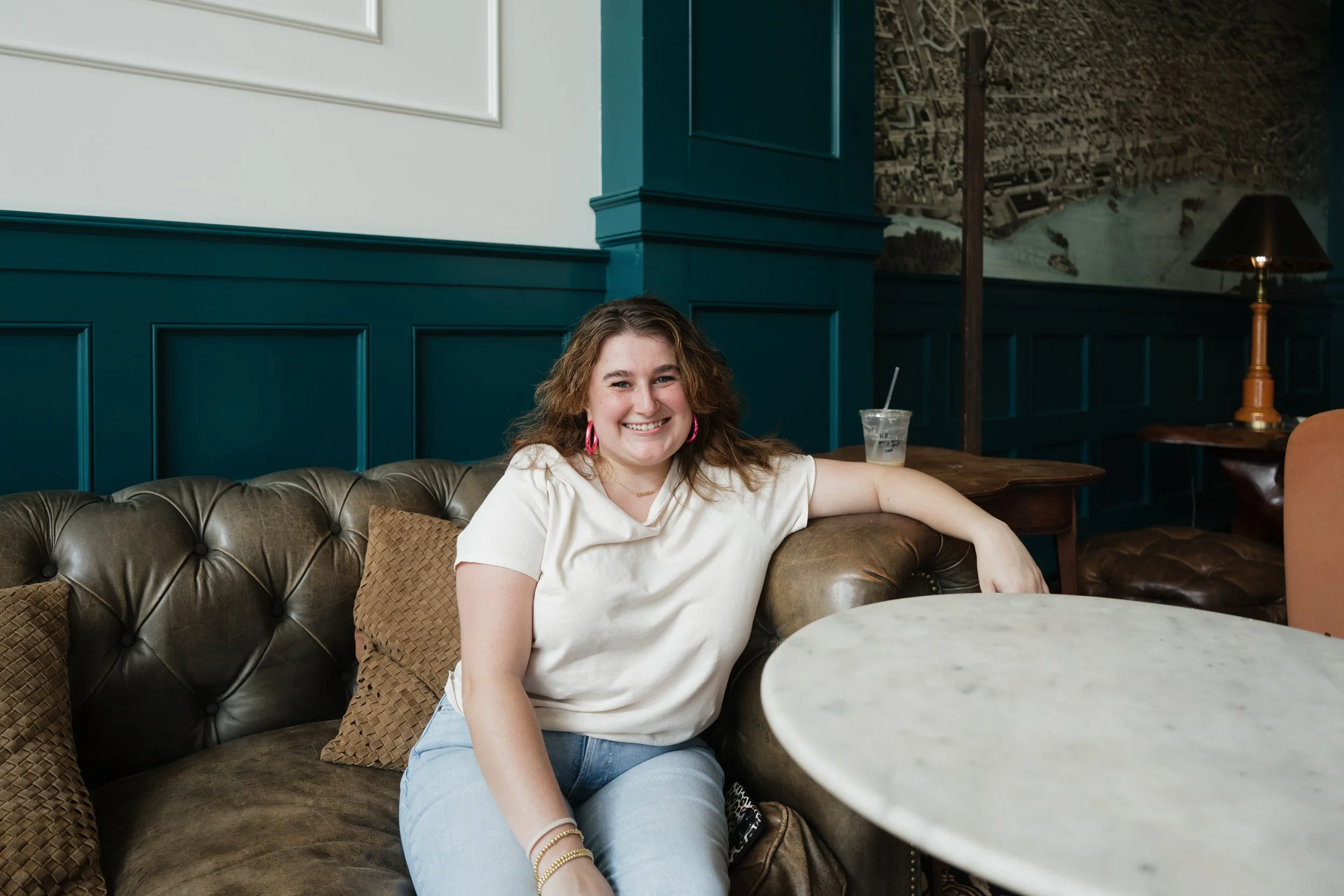 A woman with curly hair and pink earrings sitting on a brown leather couch in a stylish cafe, smiling at the camera with a drink on the table beside her.