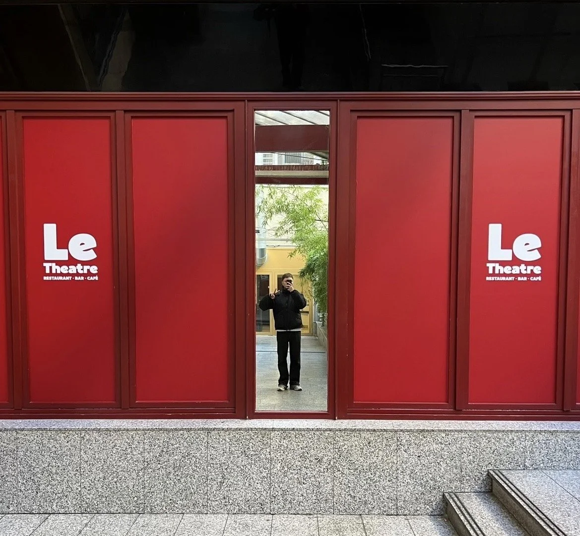 Ein Mann macht ein Selfie vor einem roten Gebäude mit dem Schriftzug 'Le Theatre', es sieht aus wie ein Spiegel, der die Person und den Hintergrund reflektiert.