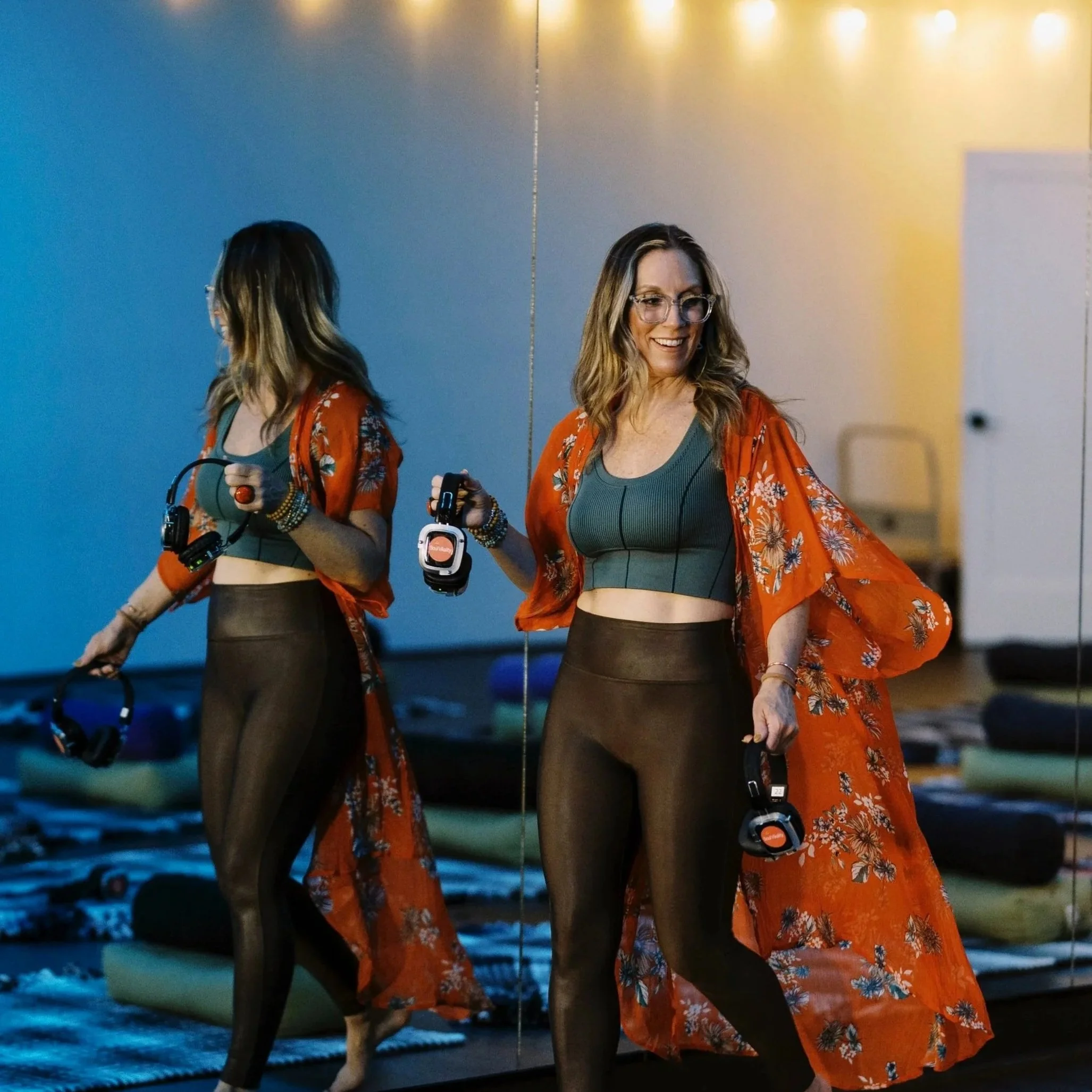 Two women standing close to each other, one facing forward with a smile and the other with her back to the camera, both in a dimly lit room with colorful lighting. One woman has glasses and wavy hair, wearing a green crop top and a red floral kimono, holding a key fob. The other woman is in a similar red floral cover-up and black leggings, with her hand holding a leash attached to a dog, whose head is partially visible at the bottom of the image.