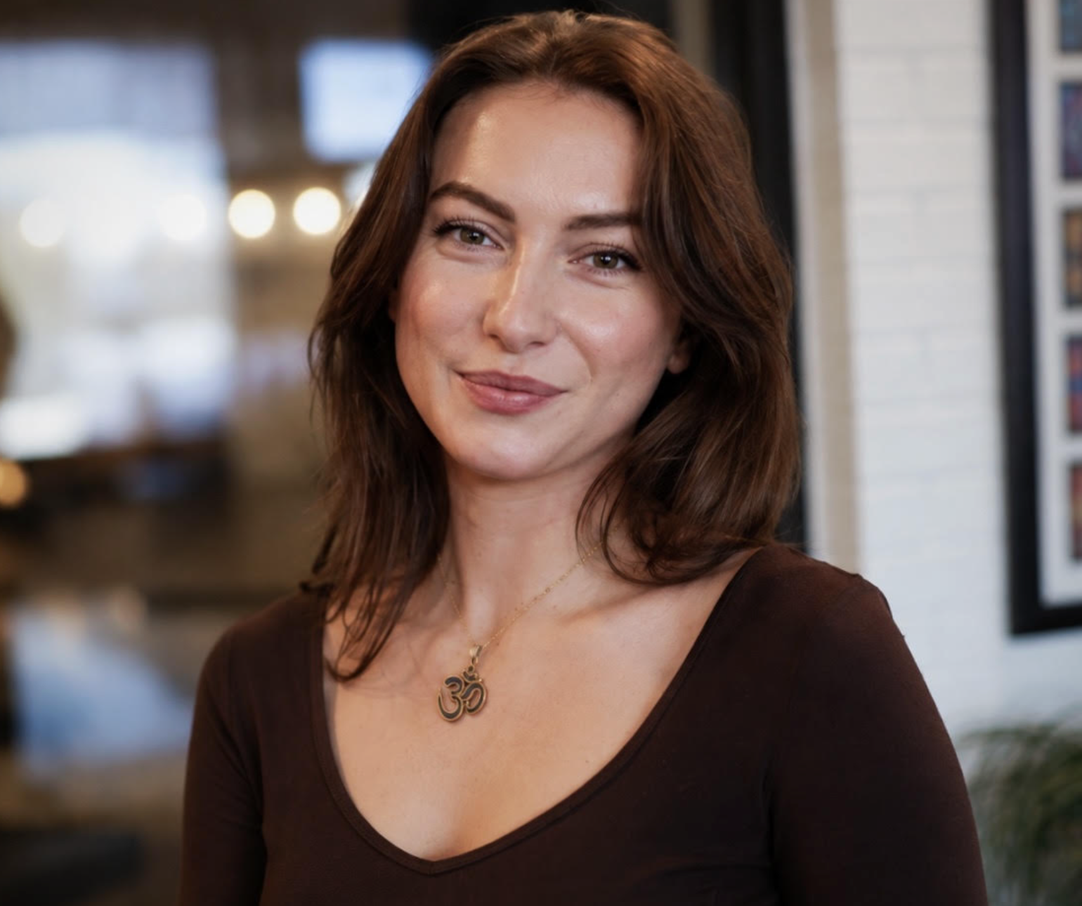 A woman with shoulder-length brown hair and fair skin, smiling softly, wearing a dark brown top and a gold necklace with an Om symbol pendant, is captured indoors with a blurred background of bright lights and interior decor.