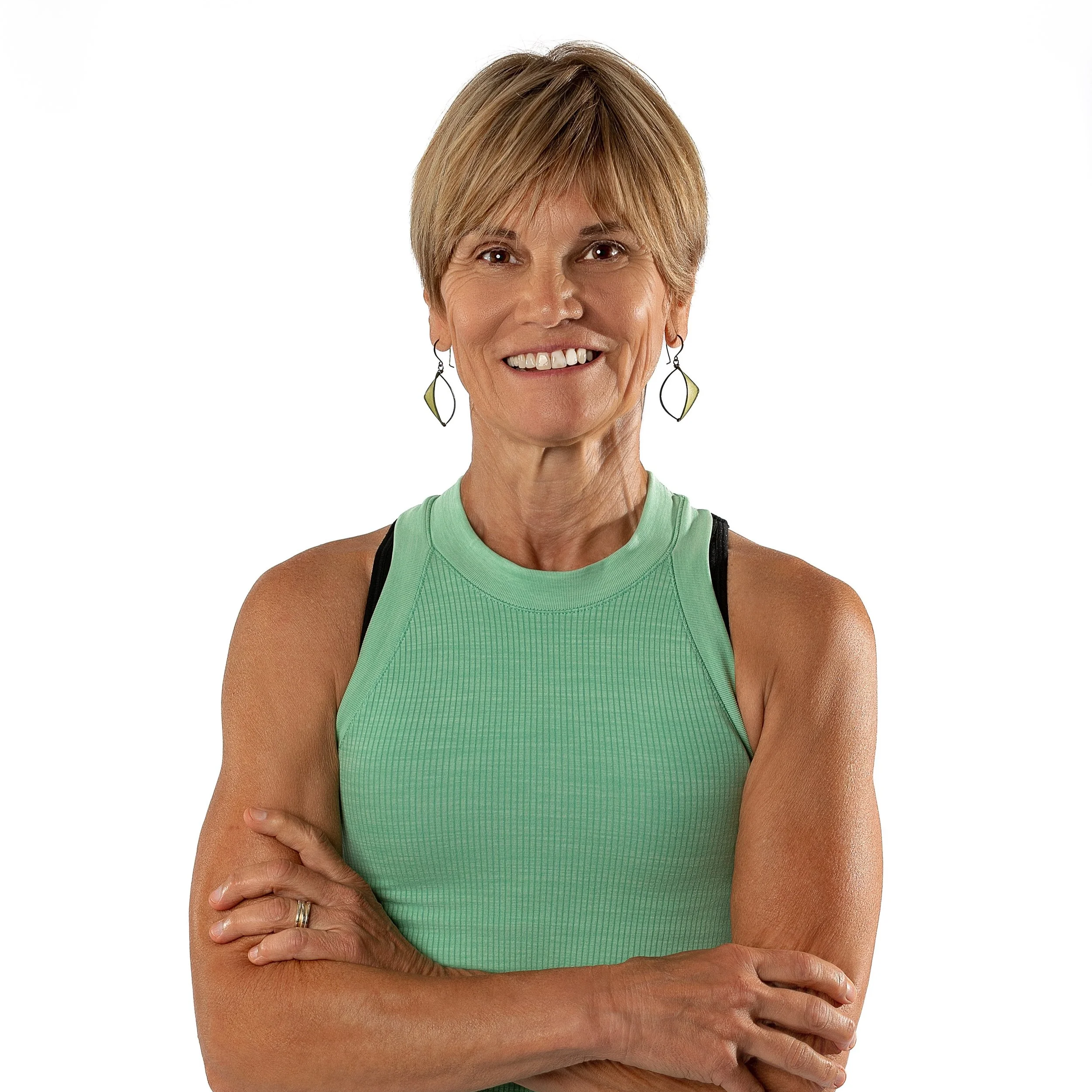 A middle-aged woman with short blonde hair smiling, wearing a light green sports top and earrings, with crossed arms, against a plain white background.