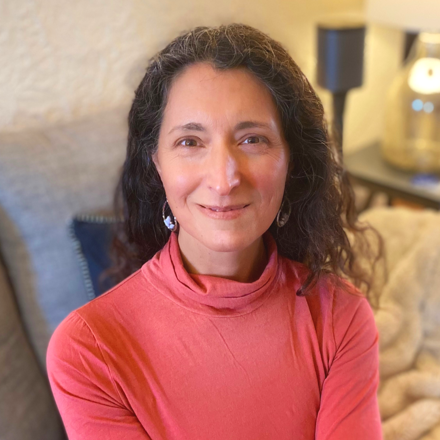 A woman with dark, curly hair wearing a pink turtleneck, smiling, sitting indoors near a beige wall and a chair with dark upholstery.