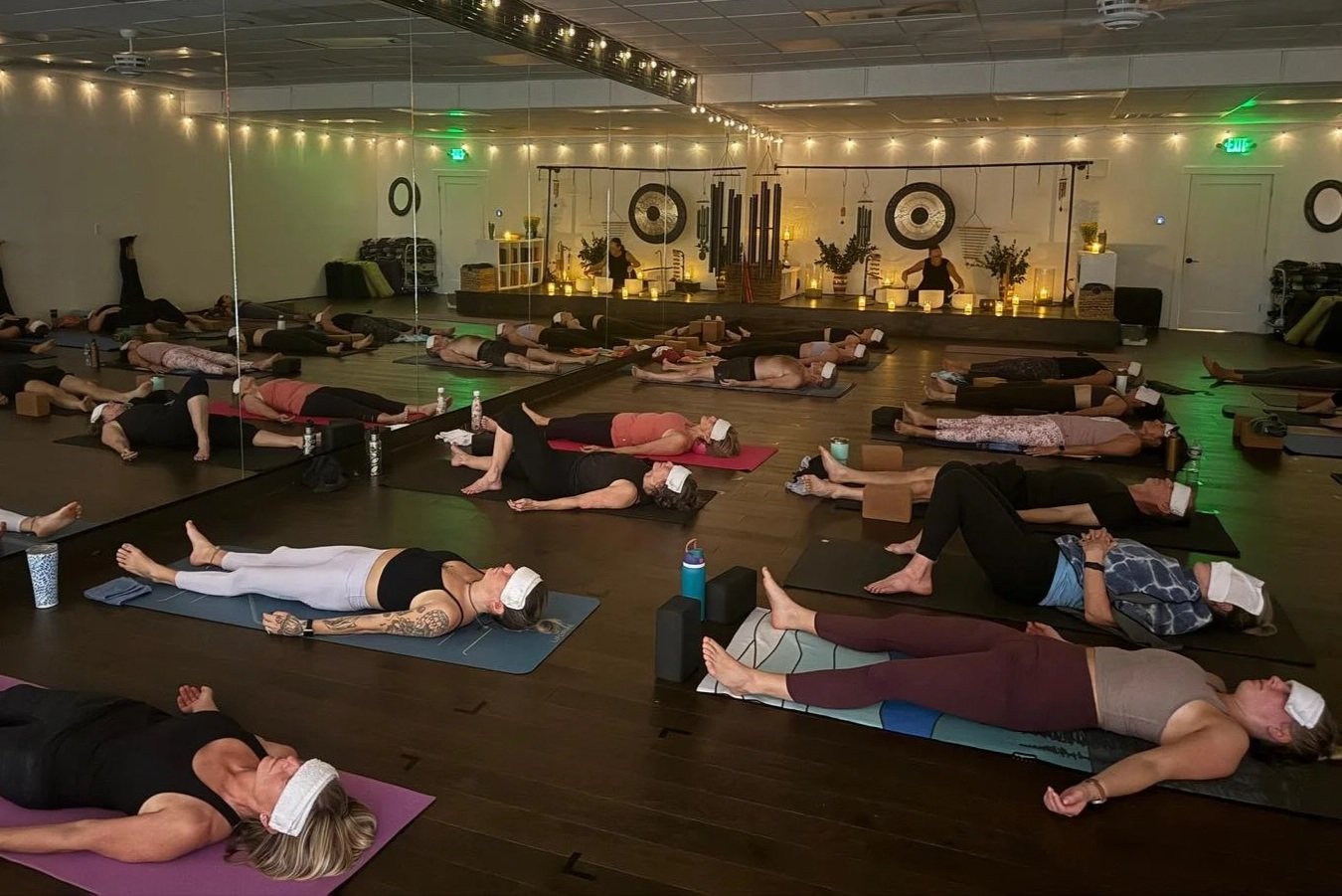 People participating in a group yoga or meditation class in a dimly lit studio, all wearing face masks and lying on yoga mats with props, with a mirrored wall and decorative elements at the front.