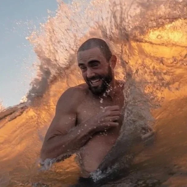 A man with a beard is swimming in the ocean, enjoying the water and smiling. The sunlight reflects on the water, creating a warm glow.