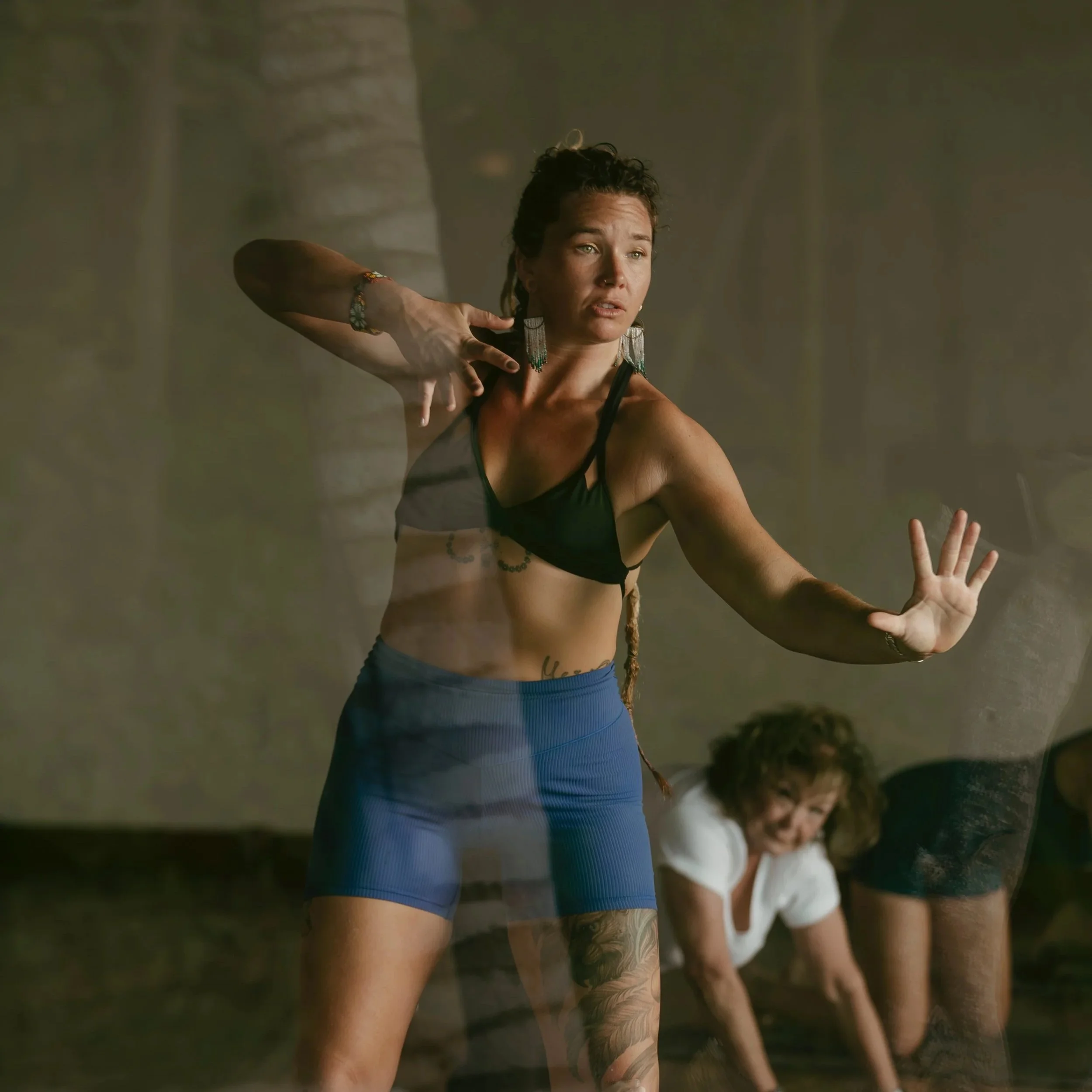 A woman with curly hair and tattoos performs a dance pose outdoors during sunset, wearing a black sports bra and blue shorts.