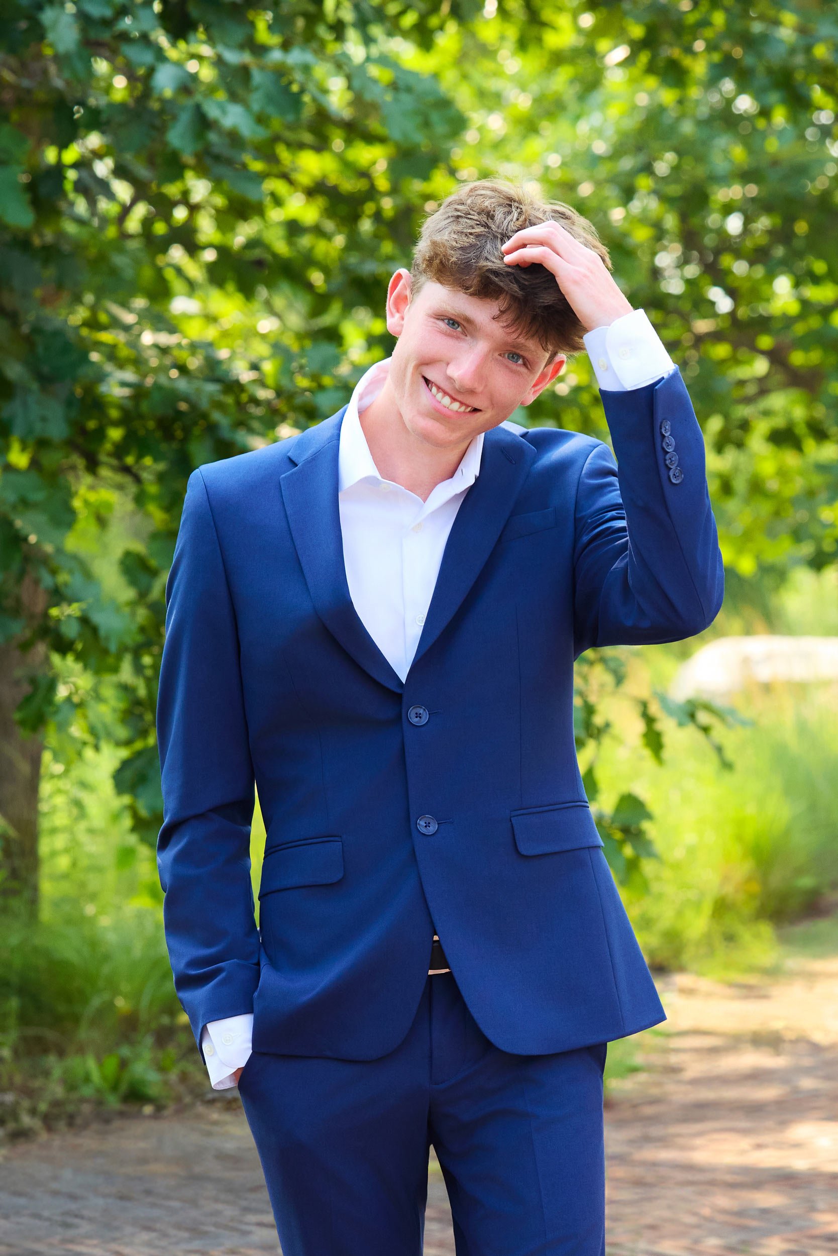 Boy wears blue suit with white shirt for high school senior picture. One hand is in pant pocket and one hand is raised, touching his hair.
